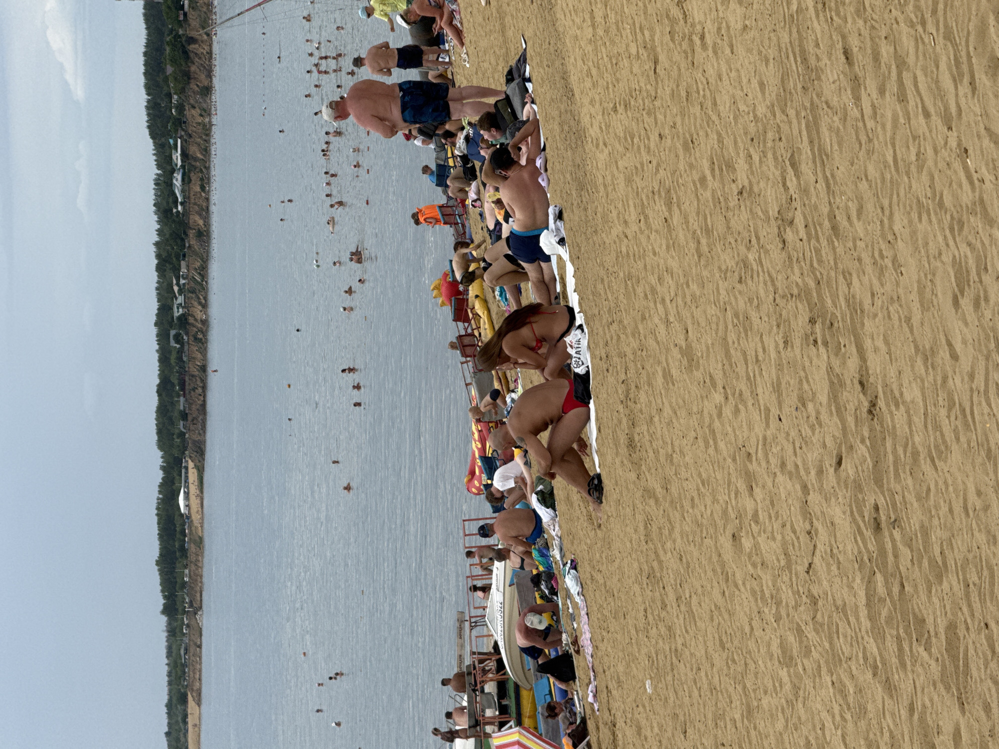 The image depicts a crowded beach on a cloudy day in Yarovoye, Russia, with the vast, calm waters of Lake Bolshoye Yarovoye extending into the distance. The foreground is dominated by a sandy beach with numerous individuals sunbathing and relaxing on towels and blankets.
In the middle ground, people are seen wading and swimming in the shallow waters of the lake, with some appearing to be engaged in conversation or playful activity. Beyond them, the far shore is lined with a dense forest and a scattering of buildings, indicative of the resort town's development. A few boats are visible moored near the shore, suggesting recreational water activities.
The scene is characterized by a leisurely atmosphere, with people enjoying the beach and water on what appears to be a typical summer day. The lighting is diffused due to the overcast sky, contributing to the soft tones of the image.