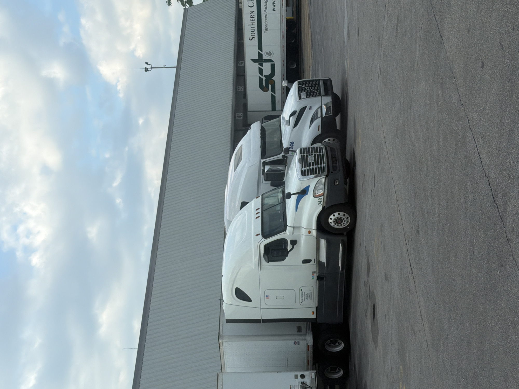 Two white semi-trucks are parked in front of a large warehouse building. The truck on the left has a white trailer and a white cab with dark gray trim. The truck on the right has a white trailer with "SCT Southern Chain" and "Paperboard Packaging" printed on it in green and black lettering, and a white cab with dark gray trim. The warehouse has a corrugated metal exterior and several loading docks marked with numbers 45, 46, 47, and 48. A light pole with two lights and an antenna is visible on top of the warehouse. The sky is overcast with light clouds. The ground is asphalt, with some cracks and water spots visible. The scene is forensic and matter-of-fact.