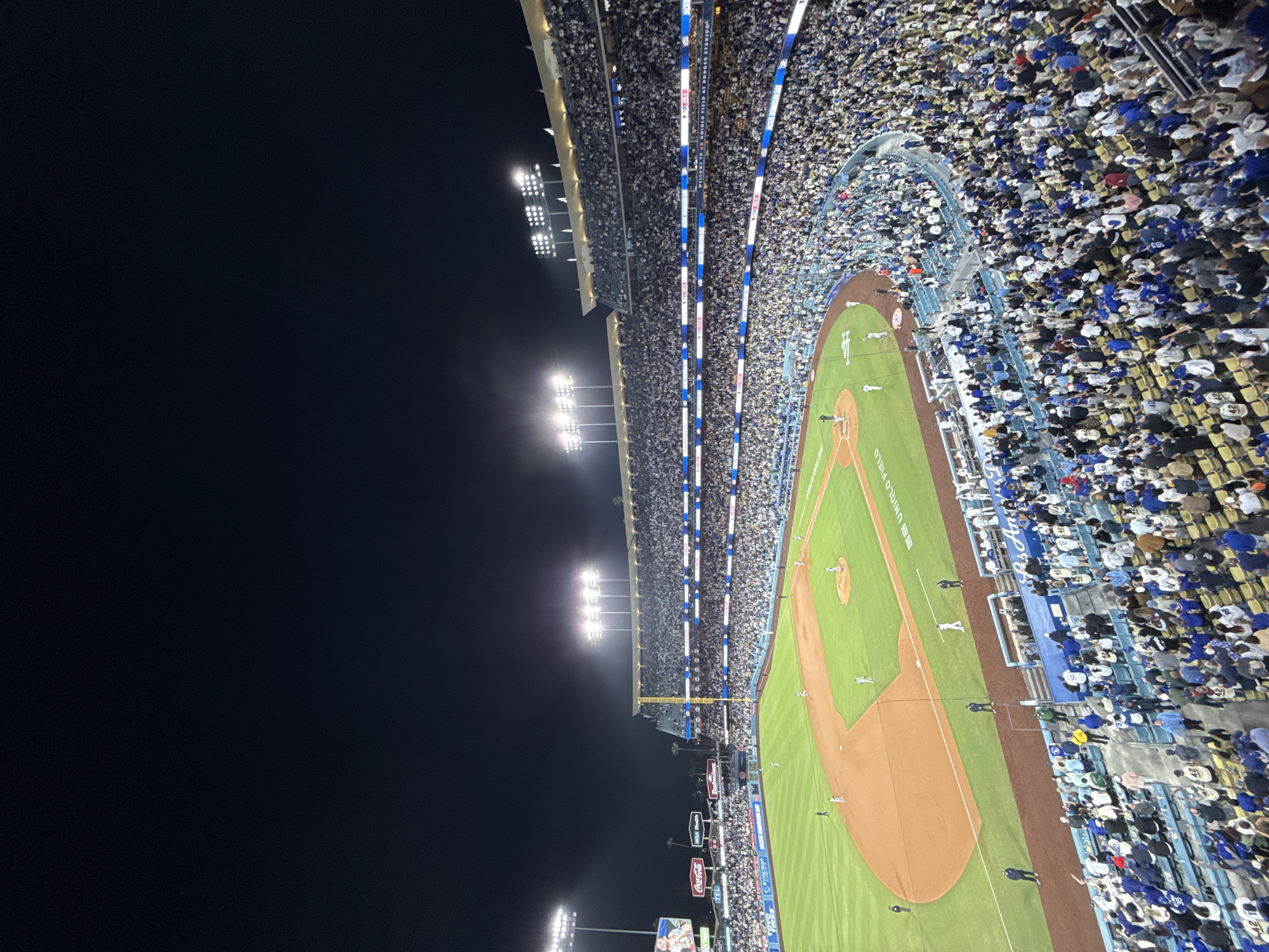 The image displays a panoramic view of a baseball game at night. The baseball field, illuminated by stadium lights, is bordered by stands packed with spectators. The fans appear to be predominantly wearing white and blue clothing. The sky is dark. In the lower left corner of the image are Coca-Cola and UCLA Health advertising signs. The location of the stadium is Los Angeles, United States of America.