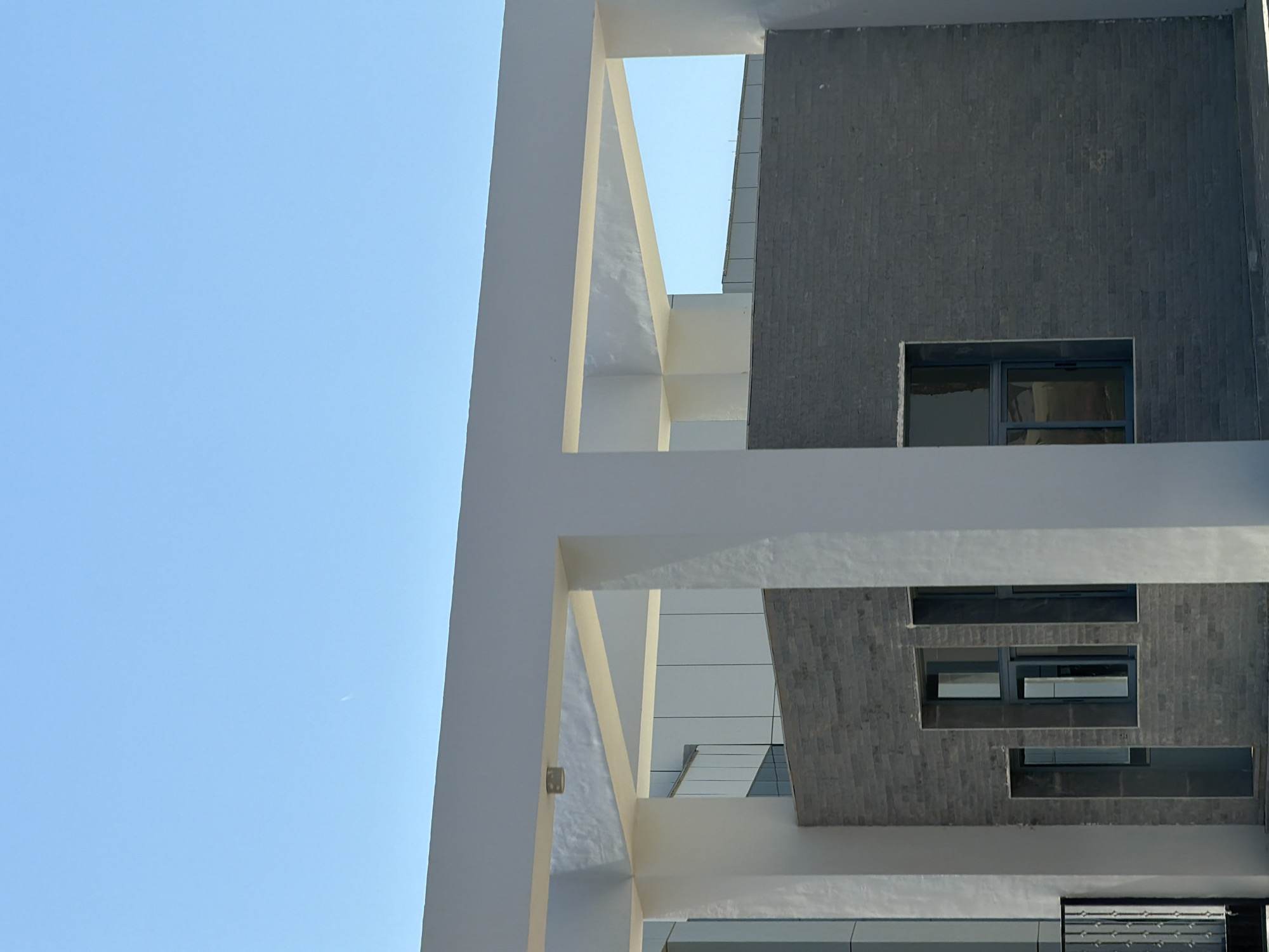 The image depicts the exterior of a modern building with a minimalist architectural style, likely in an urban or commercial setting. The structure features clean lines and a predominantly white facade, accented by a dark grey brick section. There are several windows visible, some with dark frames. A large white canopy extends across the top of the scene, supported by pillars. The sky above is clear and light blue, with a faint contrail visible. The overall impression is one of a well-maintained, contemporary building under a bright sky. The scene does not show any people or specific actions. Without further context or landmarks, it is not possible to definitively pinpoint the location within Sharm El Sheikh, Egypt.