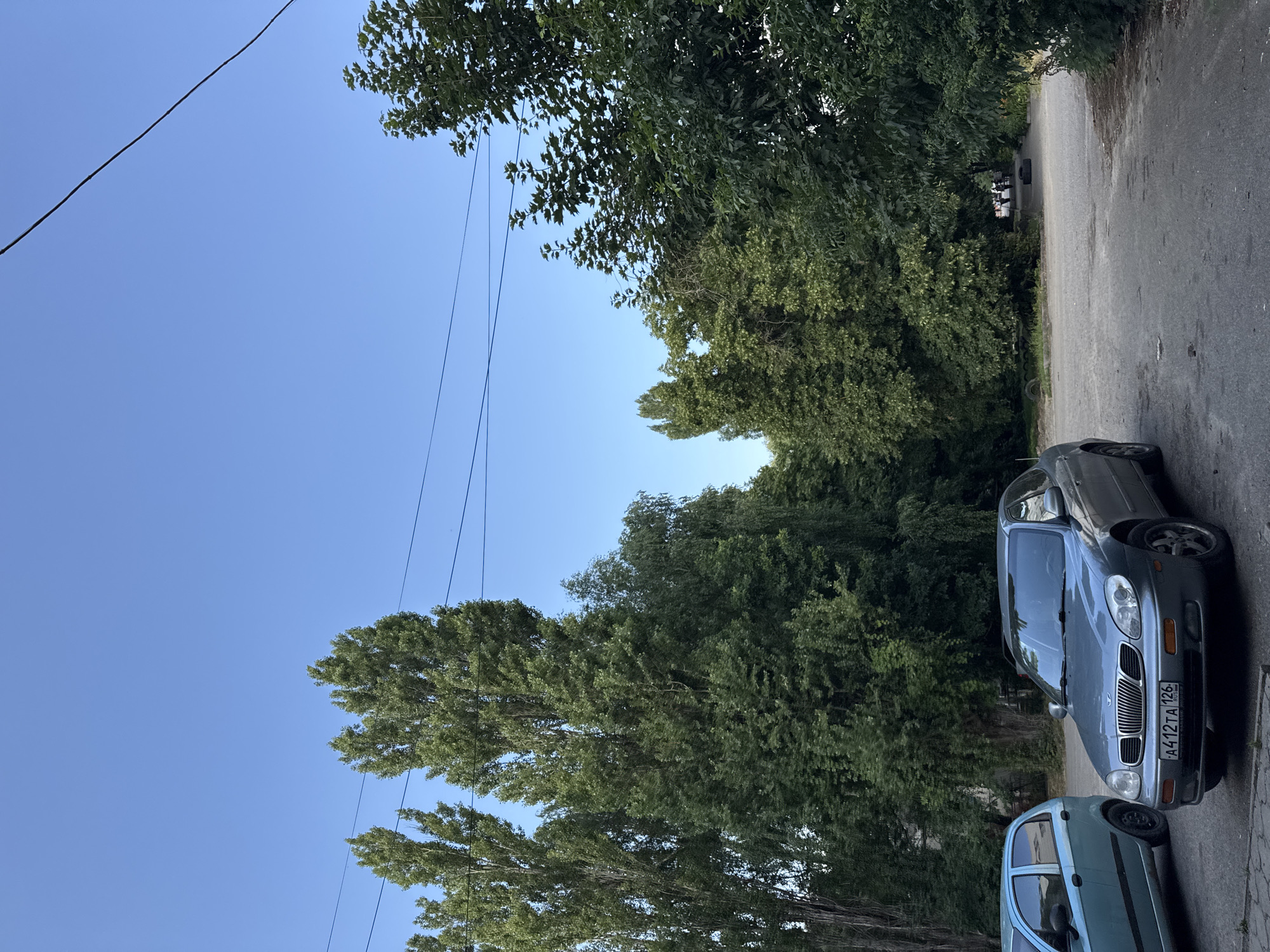 The image shows two cars parked on a paved road bordered by a dense line of trees. The sky is clear and blue, suggesting it is daytime, likely late afternoon given the soft lighting. The trees are full of green leaves, indicating a warm season. The cars are positioned diagonally on the right side of the frame, with a grey sedan in the foreground and a lighter blue car partially visible behind it. The grey sedan has a license plate that reads "A412TA 126". In the background, the trees create a lush, natural backdrop, partially obscuring a glimpse of other vehicles further down the road. The overall scene conveys a sense of everyday life in a suburban or semi-rural area. The location is identified as Verkhnerusskoye, Russia.