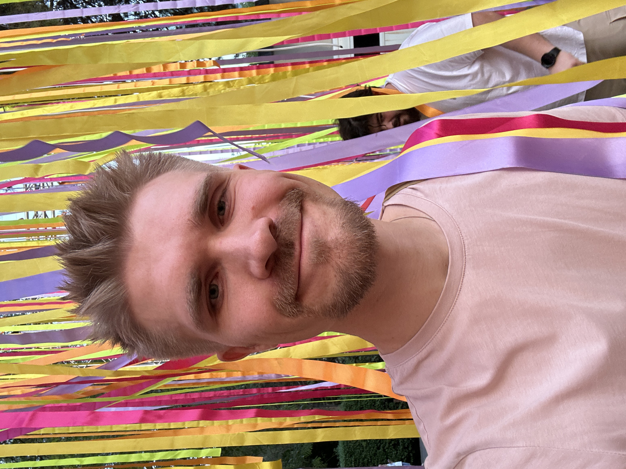 A close-up, rotated, head-and-shoulders selfie captures a man with light skin, fair hair, and a trimmed beard. He is smiling at the camera. He is wearing a light pink t-shirt. In the background are numerous colorful ribbons strung horizontally, consisting of yellow, pink, and purple. Another person is partially visible above the ribbons, wearing a white shirt and a watch. The bottom of the photo shows a glimpse of greenery. The location is not discernible as Tashkent, Uzbekistan.