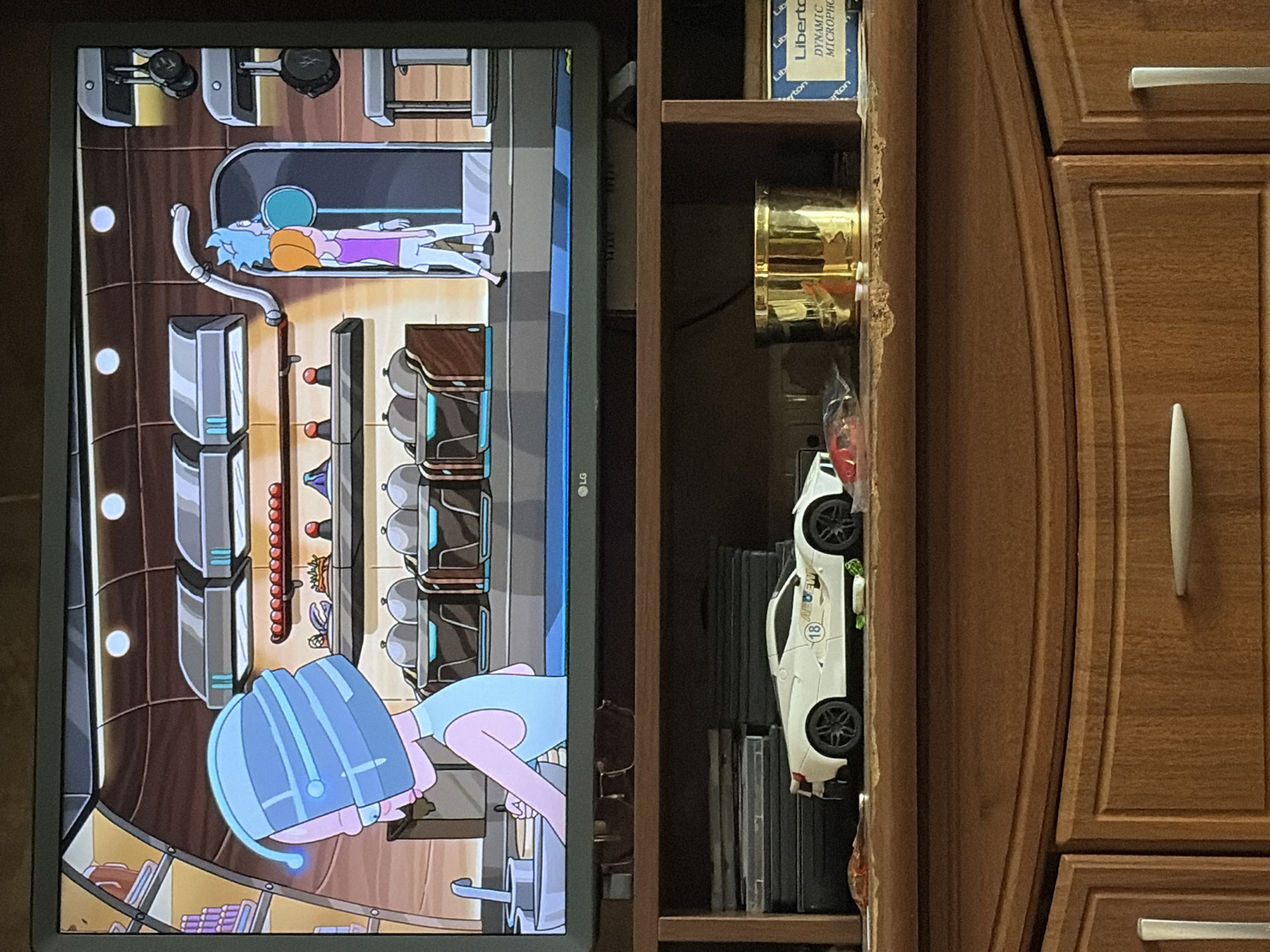 The image shows a television screen displaying a scene from the animated show "Rick and Morty." In the foreground, below the television, is a wooden cabinet with a shelf. On the shelf sits a white toy sports car with the number 18 on its side, a gold cylindrical object, and some boxes. The cabinet has wooden drawers with silver handles. The overall scene appears to be in a domestic setting. There is no information about the specific location in the city of Unknown, Unknown.