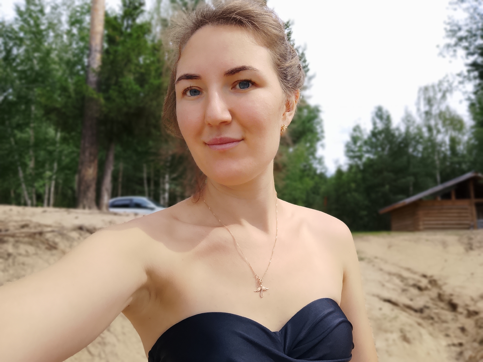 A woman with light brown hair pulled back and blue eyes is pictured in a close-up selfie. She is wearing a dark blue strapless top and a delicate gold necklace with a bird-shaped pendant. Her skin appears fair with a slight tan. The background suggests an outdoor setting with a sandy beach and trees. A blurred wooden structure, possibly a cabin or shed, is visible on the right side of the frame, along with trees in the distance. A white car is also partially visible in the background, to the left. The scene is likely set in a natural area near Noviy, Russia, given the presence of birch trees and a wooden cabin common in Russian rural landscapes.