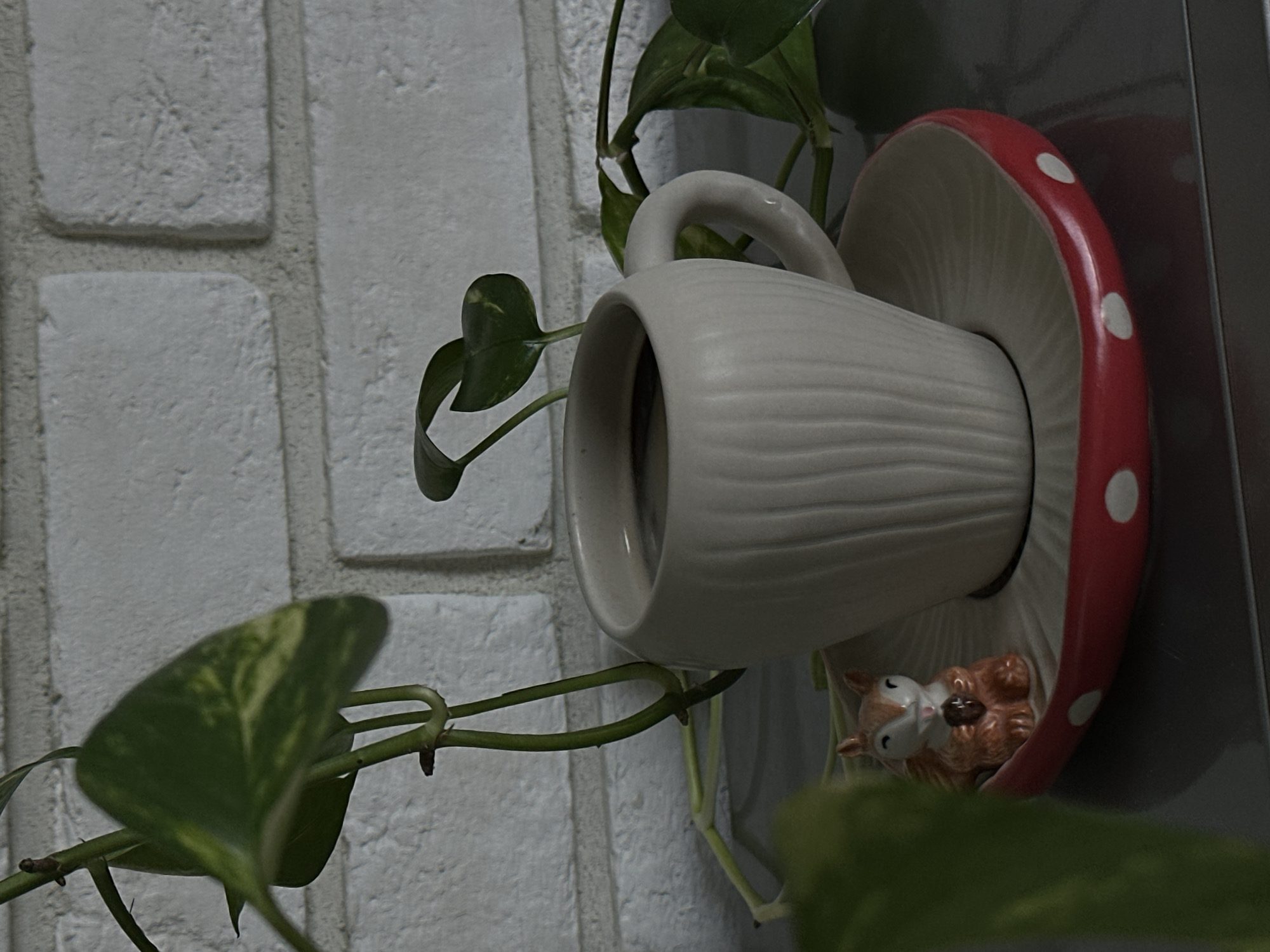 A close-up shot focuses on a ceramic mug filled with dark liquid, likely coffee, placed on a mushroom-shaped saucer with red and white polka dots. To the left of the mug, a small ceramic squirrel figurine holding an acorn sits on the saucer. The mug and saucer are on a dark, reflective surface, possibly a table. In the background, a white brick wall is visible, with green plants, including a Pothos vine, encroaching from the left and right sides of the frame. The lighting is dim, creating shadows and highlighting the textures of the objects and the brickwork. There are no people visible in the image. The scene is set indoors, with no specific geographic indicators to place it in Khimki, Russia, or any other city.