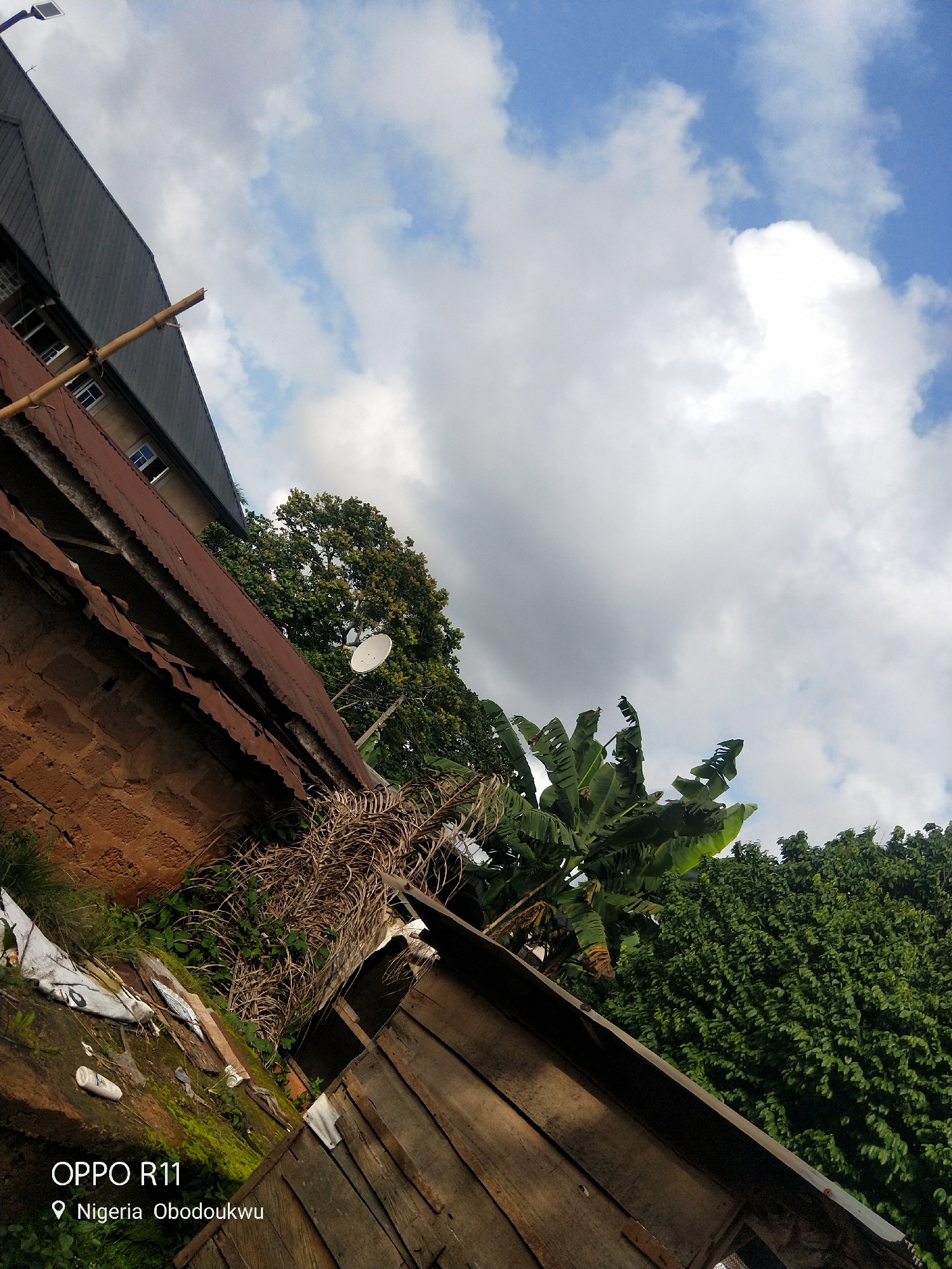 The image shows an outdoor scene under a bright, cloudy sky. In the foreground, there is a weathered wooden structure with a corrugated metal roof. To the left, a brick wall is partially visible, covered in moss and overgrown with dry, dead vegetation and some greenery. A white plastic bottle and other debris are scattered on the ground.
Further into the scene, behind the wooden structure and brick wall, are lush green trees, including banana trees with large, distinctive leaves. A satellite dish is mounted on a pole, facing upwards. In the background, a multi-story building with a dark, sloping roof and visible windows can be seen. A long, wooden pole extends diagonally across the building and into the sky. The overall impression is of a somewhat rural or developing area.
The weather appears to be fair with scattered clouds and bright daylight, suggesting it is daytime. The location is identified by visible text at the bottom left of the image: "OPPO R11" and "Nigeria Obodoukwu".