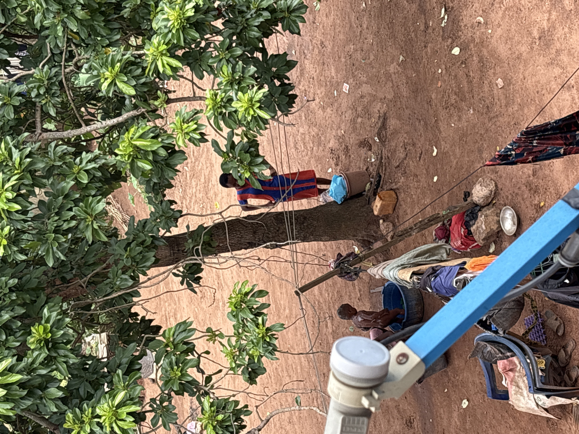 The image captures an outdoor scene in a seemingly rural or underdeveloped area in Onitsha, Nigeria. A large tree with abundant green foliage dominates the center of the frame, its branches extending across the top portion of the image. The ground is covered in reddish-brown earth, with scattered leaves and debris.
In the mid-ground, a young child, appearing to be bathing, stands in a blue basin filled with water. The child's skin is visibly wet. Nearby, an adult, possibly a woman, stands with hands on hips, wearing a blue and red striped dress. She is positioned near the base of the tree. Several items of clothing are draped over a wooden pole, suggesting laundry is being done or has been recently washed. Various buckets and containers are scattered around the area.
In the foreground, a satellite dish receiver is prominently visible, its blue arm extending diagonally across the image. To the left of the satellite dish, a blue plastic chair is overturned, with laundry and other items placed on it. Flip-flops are visible on the ground near the chair.
The overall impression is of a simple, outdoor living space, likely within a residential area, where daily chores are being carried out under the shade of a large tree. The presence of the satellite dish suggests some level of modern infrastructure, contrasting with the rustic environment.