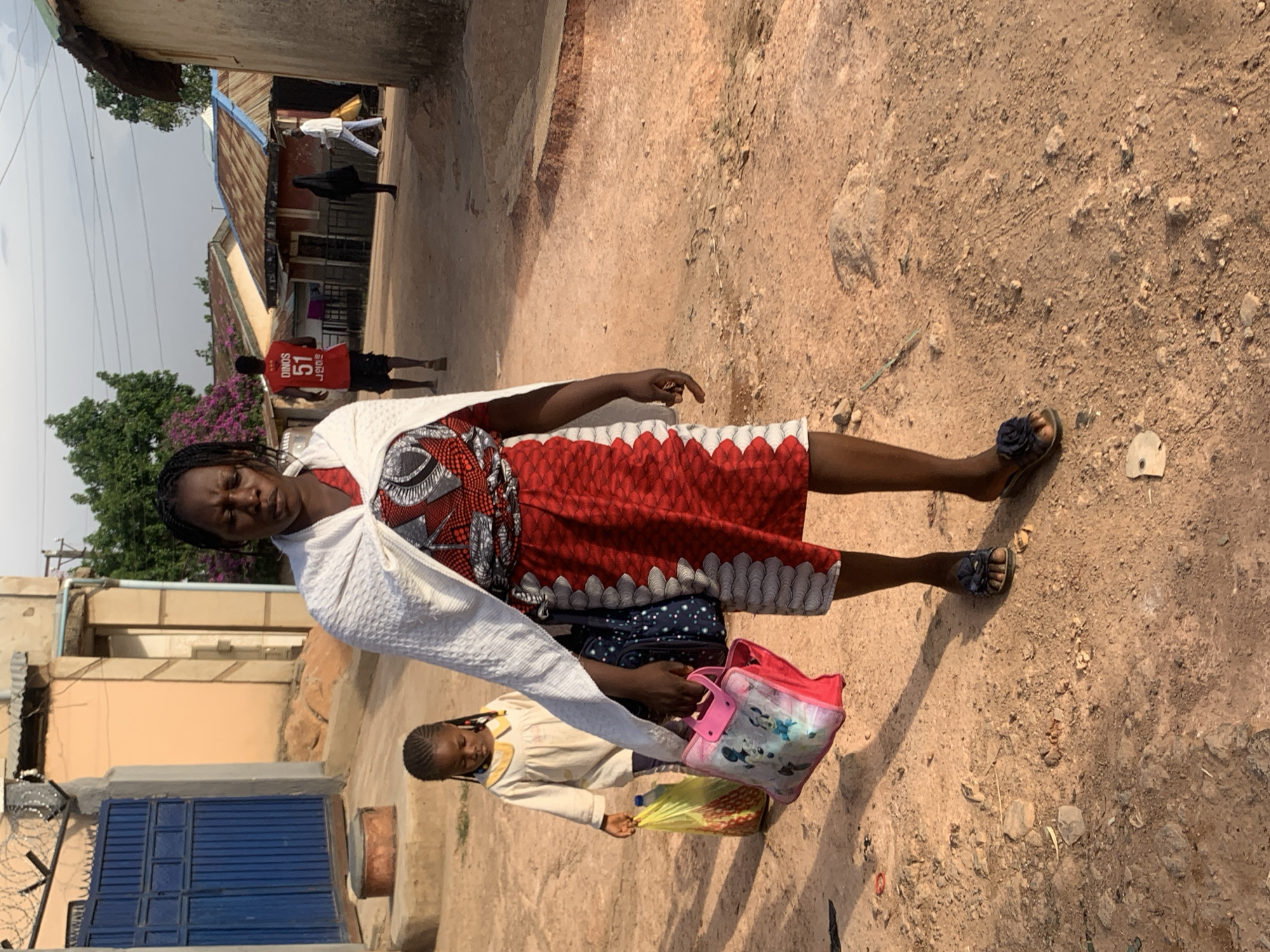 A woman wearing a red and white patterned dress, a white shawl, and sandals stands on a dirt road in Jos, Nigeria. She is carrying a pink and white tote bag in her left hand. Beside her is a young girl in a tan outfit carrying a yellow plastic bag. Behind them, a man in a red jersey with the number 51 walks away. In the background are buildings with corrugated metal roofs. The road surface is a mix of dirt and gravel.