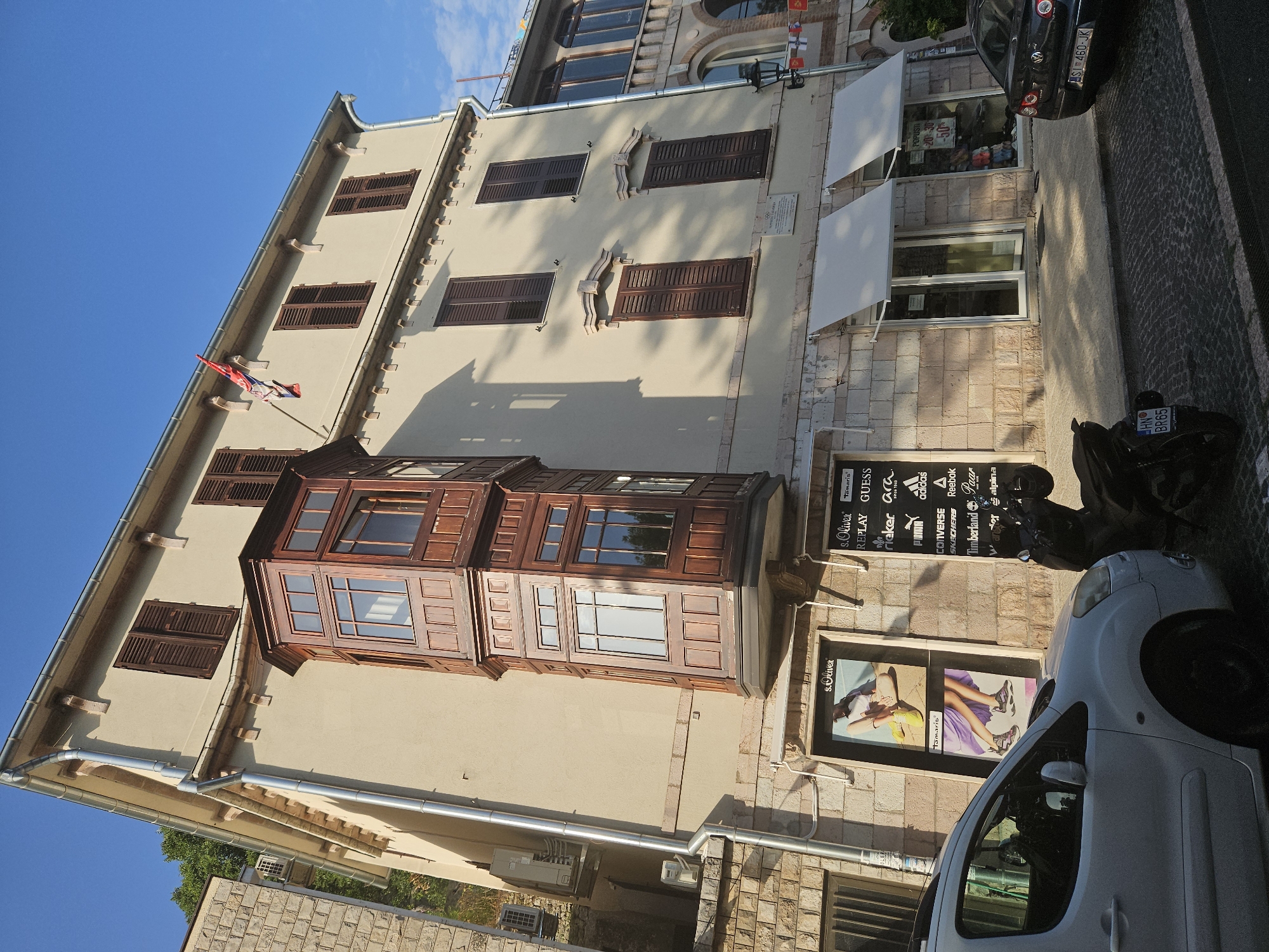 The image shows a sunlit street scene in Herceg Novi, Montenegro, featuring a building with a prominent wooden bay window. The building, painted a light yellow, has dark brown shutters on its upper-story windows and is adorned with a flag on the top right. The lower level of the building houses several retail storefronts, including one displaying advertisements for fashion brands like s.Oliver and Tamaris, and another featuring a list of sportswear and shoe brands such as Replay, Guess, Adidas, and Puma. A white van is parked on the left side of the street, partially obscuring the storefronts. To the right of the van, a black motorcycle is parked. A black sedan is visible further down the street on the right. The street is paved with cobblestones, and a shadowed walkway runs in front of the shops. The sky is a clear blue.