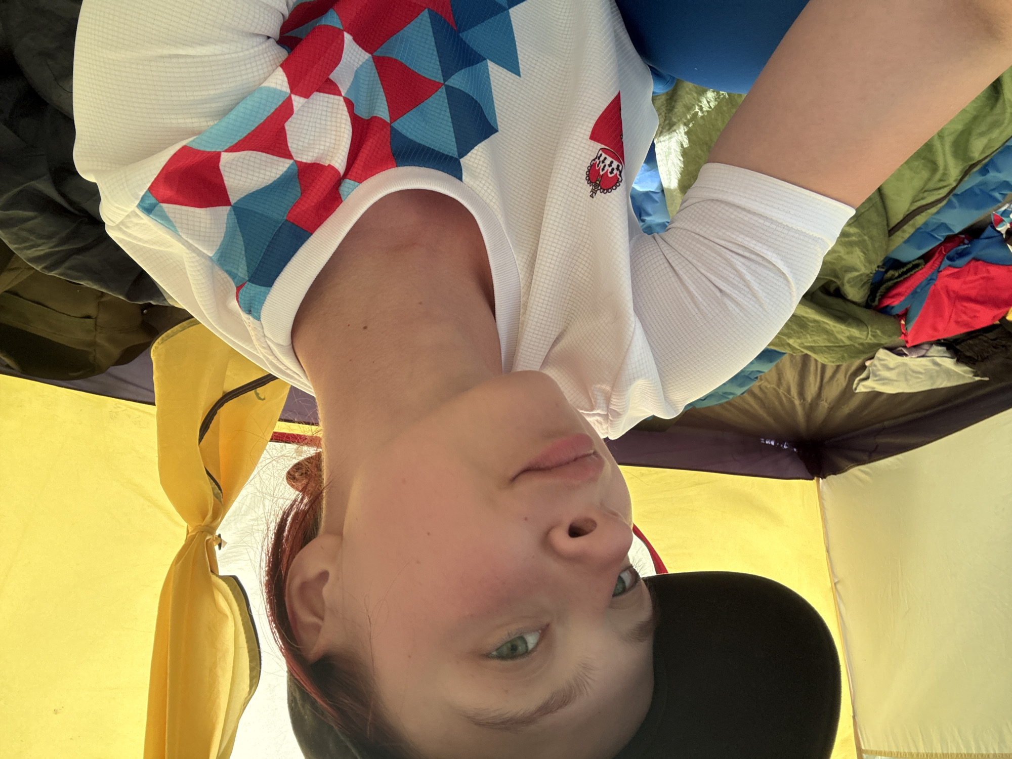 A person is lying inside a tent. The tent's interior walls are pale yellow and purple. The person is wearing a white athletic shirt with red and blue geometric designs on the upper chest and shoulders, along with a small emblem on the right side of the chest. They are also wearing a black baseball cap. The ground around the person is covered with a mixture of dark and light camping equipment, including a sleeping bag, various cloths, and tent equipment. The photo appears to have been taken from a low angle inside the tent. There is no indication of the city of Khvalynsk, Russia.