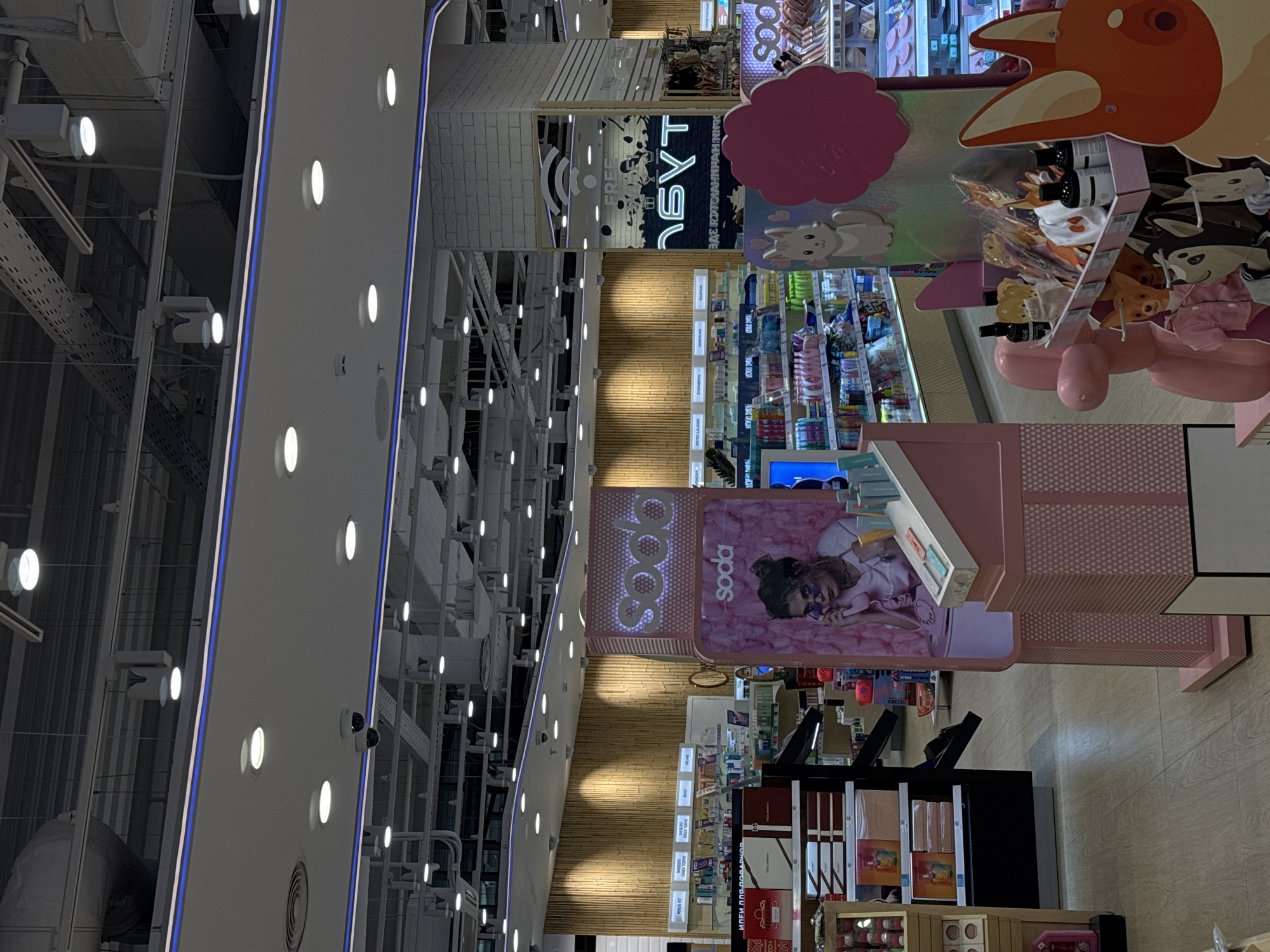 The image shows the interior of what appears to be a retail store, likely a beauty or cosmetics store. The store has a modern design with bright lighting and a high ceiling. The ceiling features a complex network of pipes and lighting fixtures. The walls are white with blue accents. There are several aisles of products on display, including cosmetics, skincare, and fragrances. The products are arranged on shelves and displays. A large, colorful display with a cartoon-like design is visible near the center of the image. The store is located in an unknown city, as the location information is not provided.
