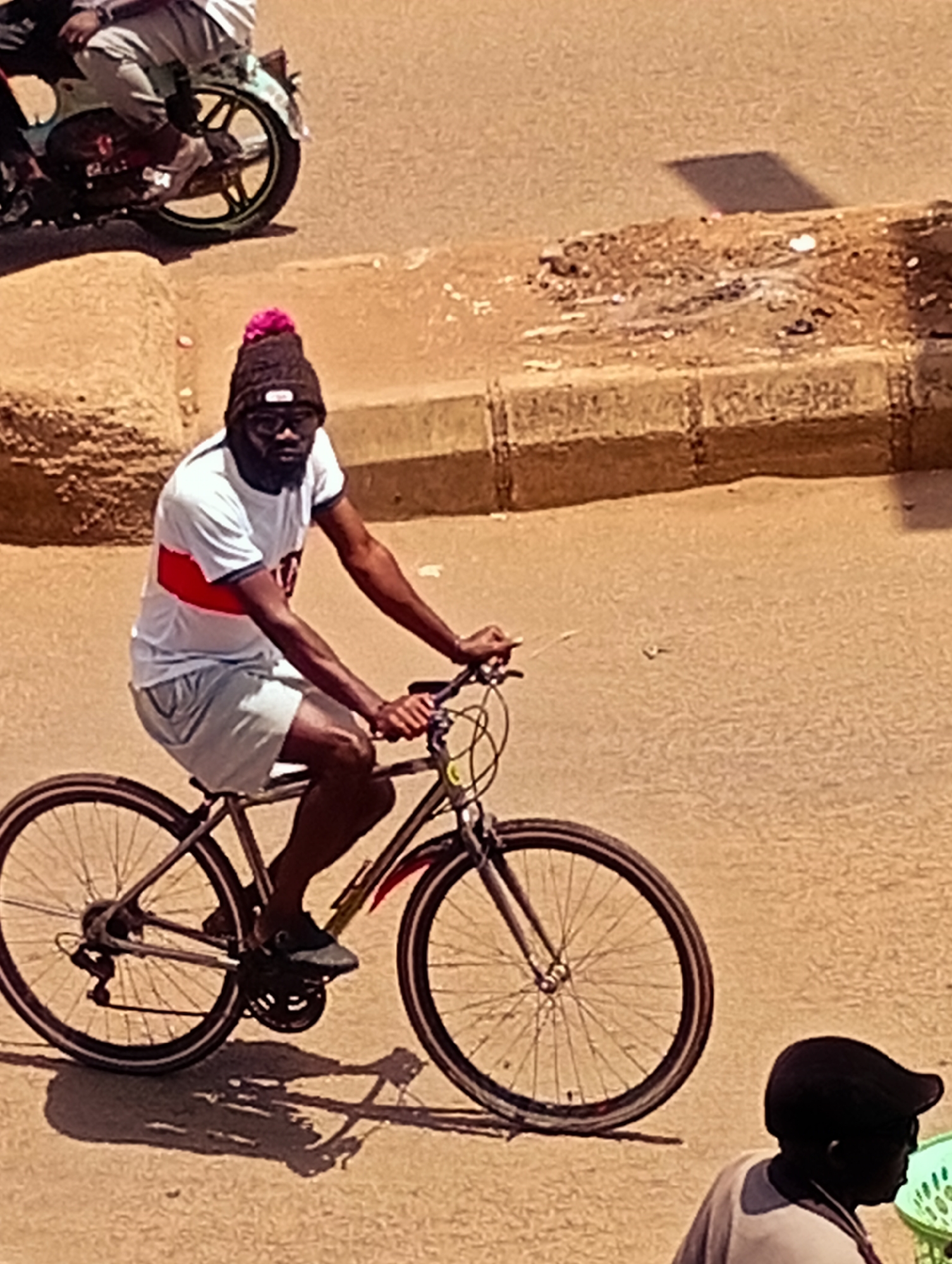 In Bukuru, Nigeria, a man wearing a black beanie with a pink pom-pom and glasses is riding a bicycle on a paved road. He is wearing a white t-shirt with a red stripe across the chest and light-colored shorts. The bicycle is silver with black wheels and tires. In the background, another person is partially visible on a motorcycle. The environment appears to be a street or roadside with a curb and rough ground. The lighting suggests it is daytime, casting shadows on the ground.