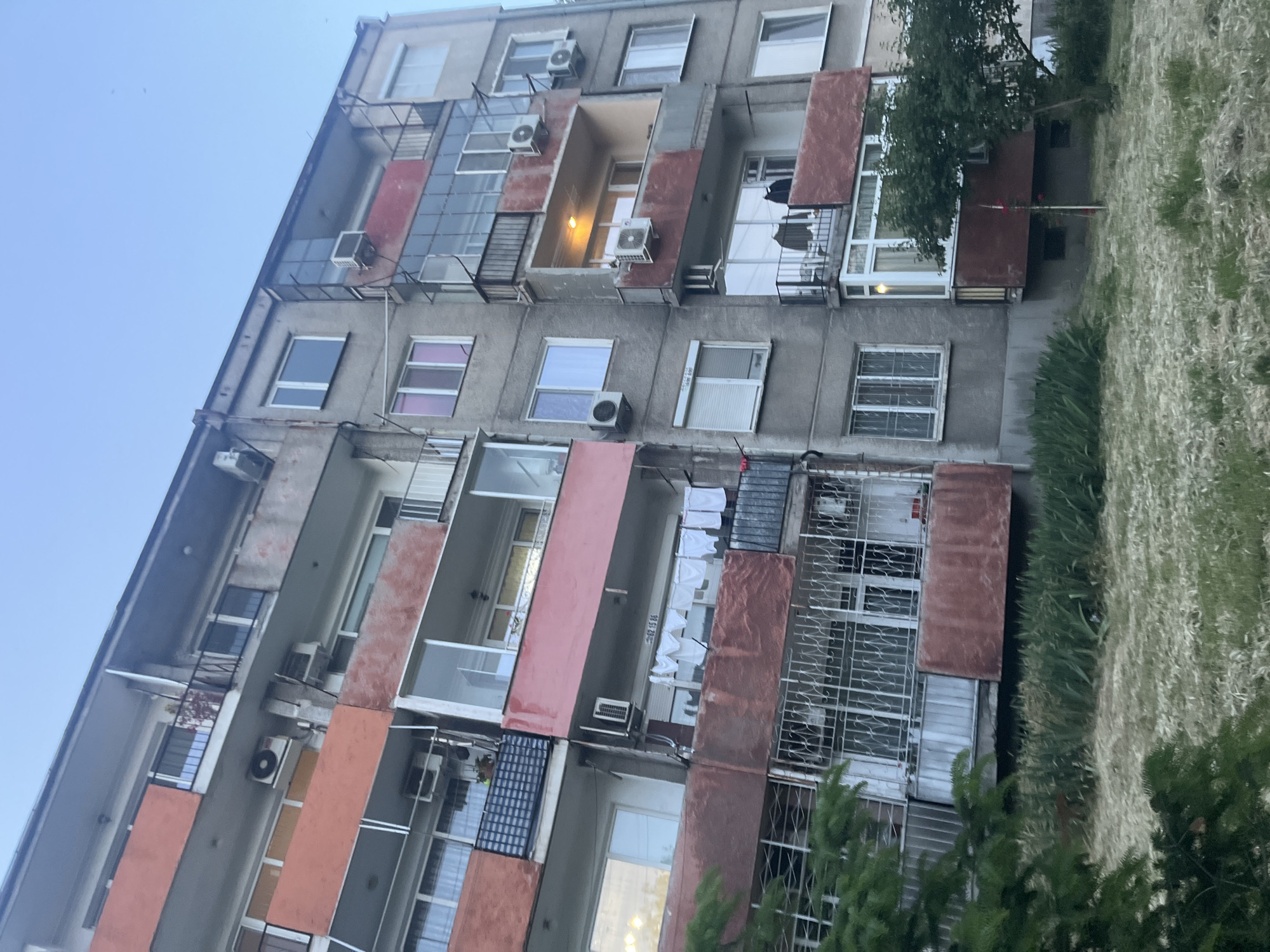 The image shows a multi-story residential building in Plovdiv, Bulgaria. The building is made of concrete and has several balconies, some of which are enclosed. The balconies are painted in various shades of red and orange. Air conditioning units are visible on the exterior walls. There are windows of different sizes and shapes, some with curtains or blinds. Laundry is hung to dry on one of the balconies. The building is surrounded by greenery, including trees and grass. The sky is a pale blue color.