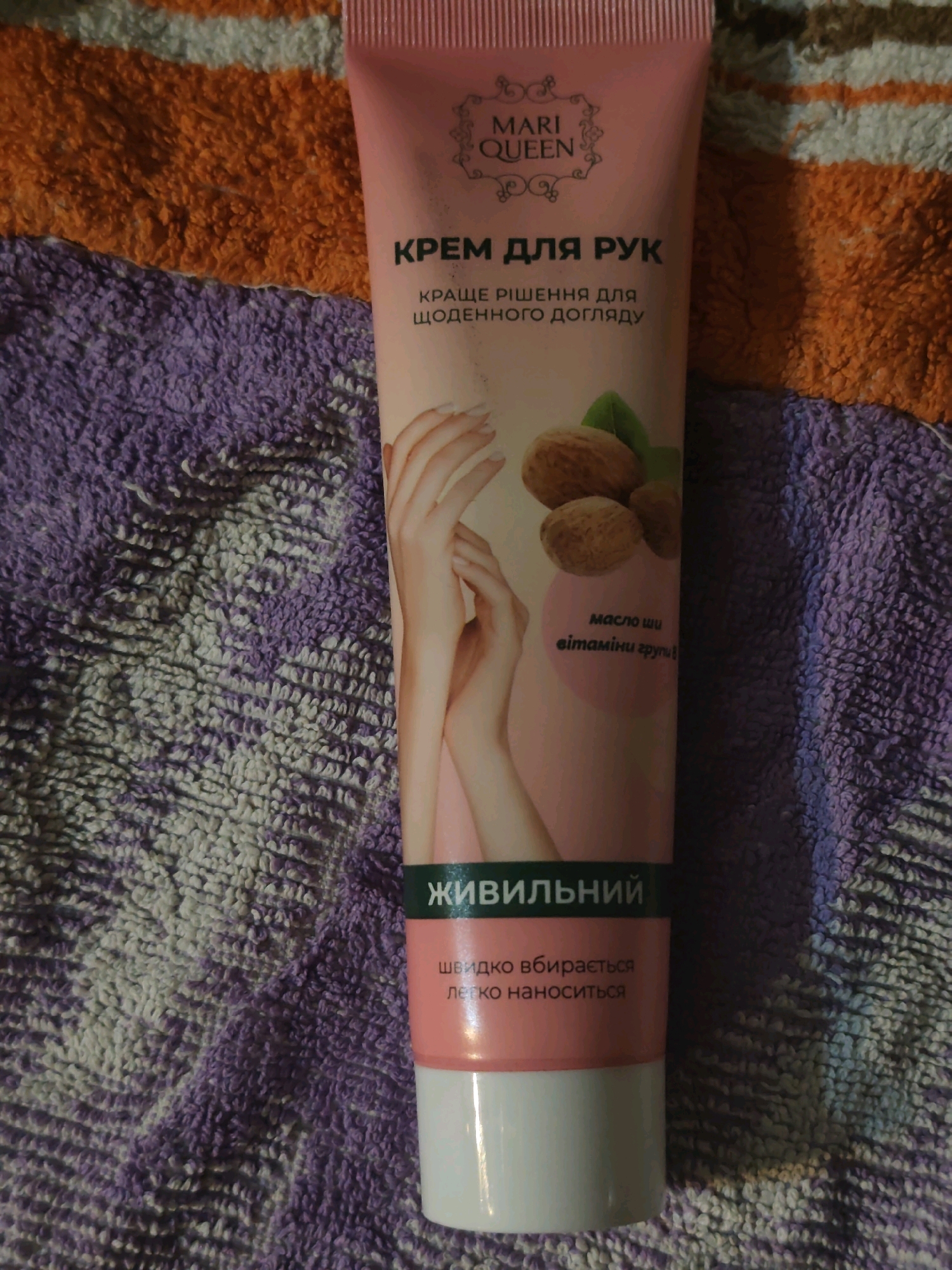 A pink tube of hand cream by Mari Queen is shown. The tube is lying on a textured surface, with parts of orange and purple fabric visible. The hand cream packaging features text in Ukrainian, indicating it is "Cream for hands" and boasts "better solution for daily care." Images of walnuts and leaves are also present on the packaging. A green band across the tube reads "Lively." Below this, smaller text states "quickly absorbs and is easy to apply." The tube has a white cap at the bottom.