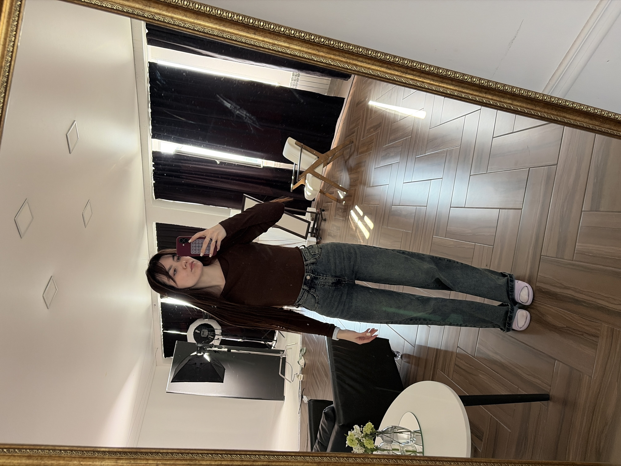 A young woman stands in front of a mirror in a room. She is taking a selfie with a smartphone. She is wearing a brown long-sleeved shirt, jeans, and light purple shoes. The room has a wooden floor with a herringbone pattern. Behind her, there is a table with a vase of flowers, a black chair, and a ring light with a softbox for photography. The background includes a window with partially obscured blinds, a folding chair, and a white wall with small square decorations. Based on visual cues, there's no readily available information to confirm the location as Chaikovsky, Russia.