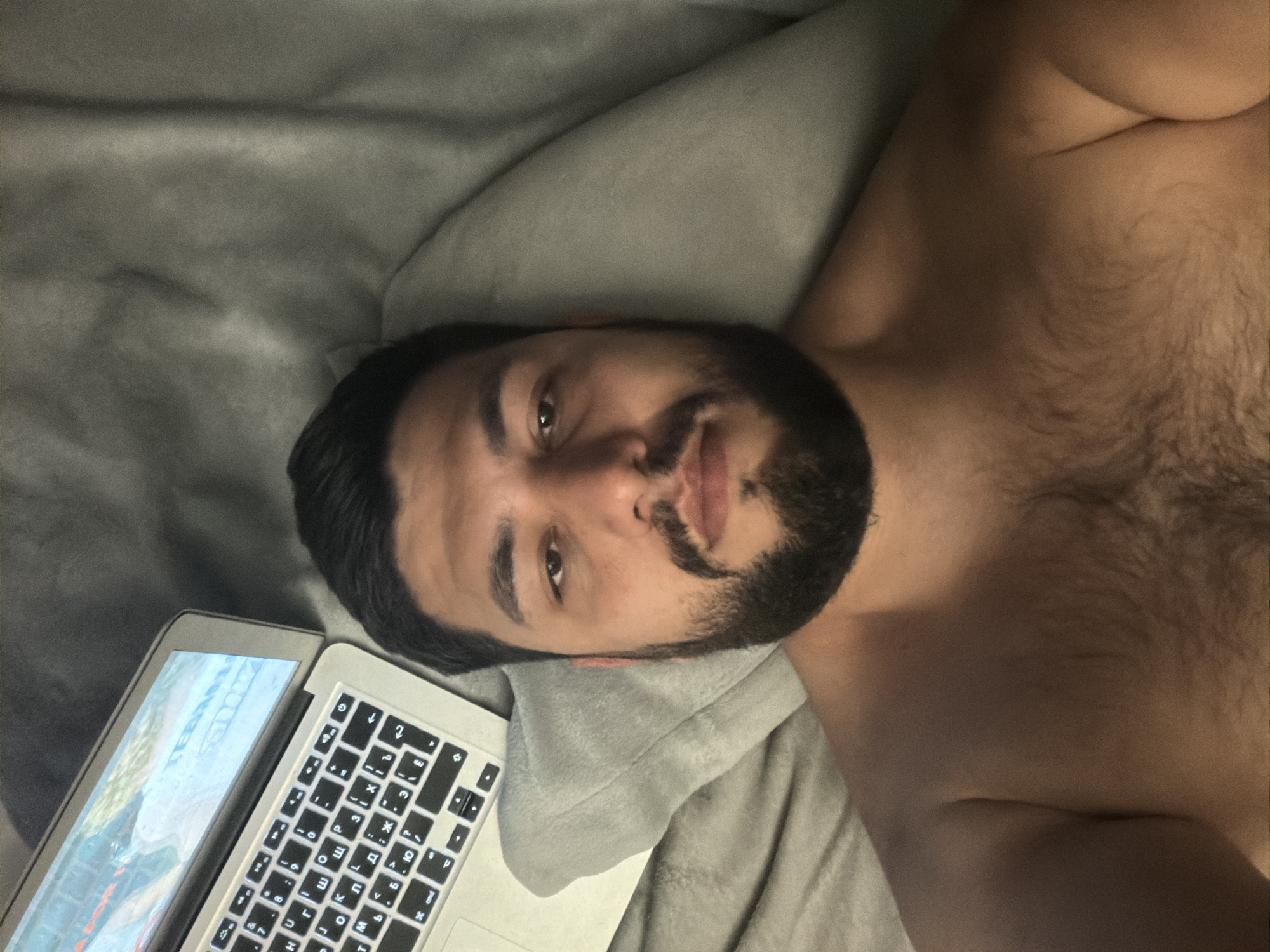 The image shows a bare-chested man lying on a gray blanket with a laptop to his side. The laptop screen is visible, displaying what appears to be a game. The man has dark hair, a beard, and is looking at the camera. The scene is indoors. There is no indication of the location being in Krasnoyarsk, Russia.