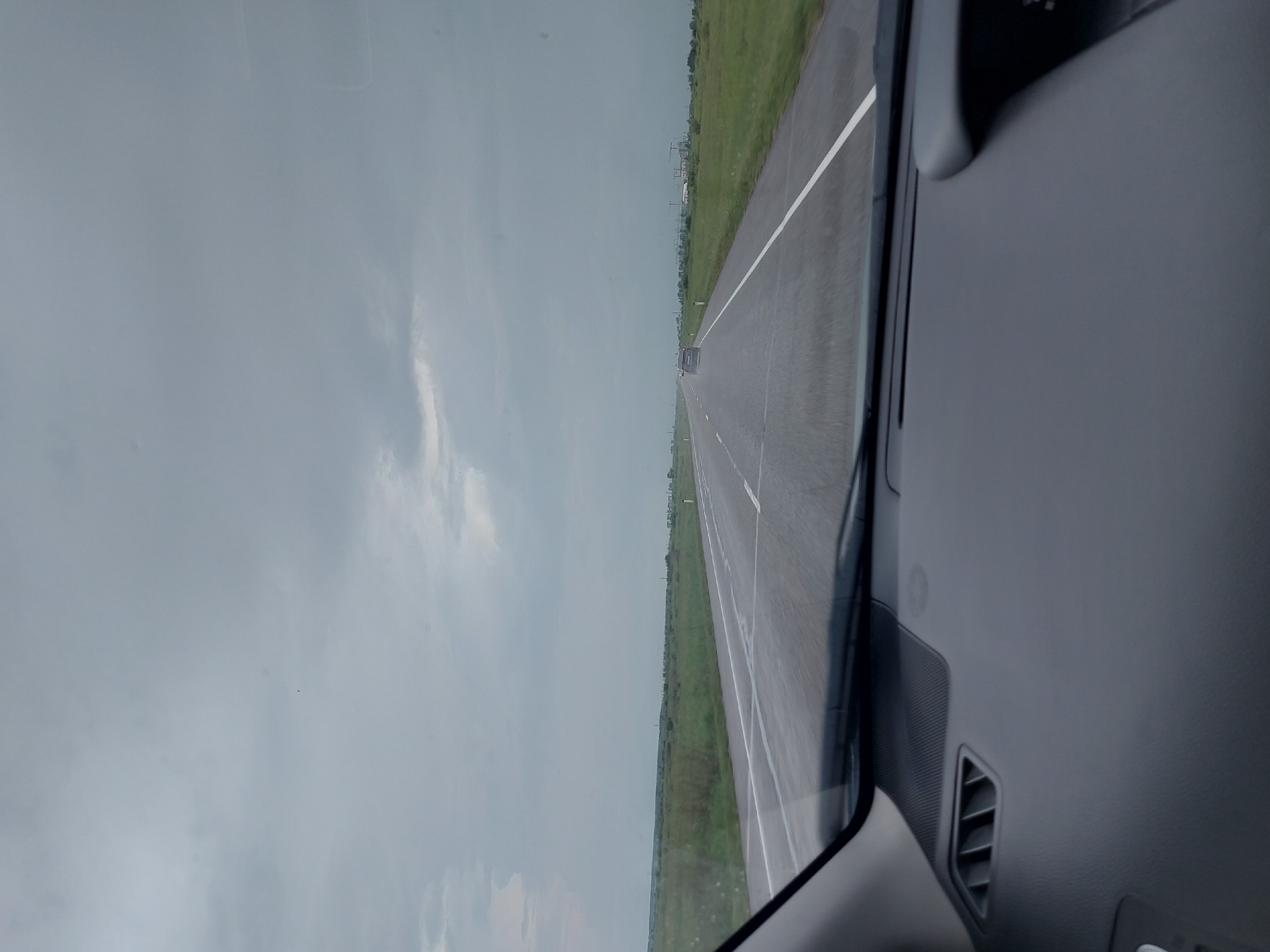 The image, taken from inside a vehicle, shows a desolate highway under a cloudy, overcast sky. The road is wet, suggesting recent rain or damp conditions. The surrounding landscape is flat and consists of green fields with sparse vegetation, extending to the horizon. The highway stretches into the distance, with white lines marking the lanes. A dark-colored car is visible on the road ahead, heading in the same direction. In the far distance, on the right side of the road, there are some buildings under construction, possibly cranes and structures. The interior of the vehicle's dashboard is visible in the foreground, with an air vent on the left. The overall impression is of a journey through a sparsely populated rural area. The scene does not depict any specific landmarks or features that would clearly place it in Novyi Byt, Russia, but the visual elements are consistent with a typical highway in a rural setting.