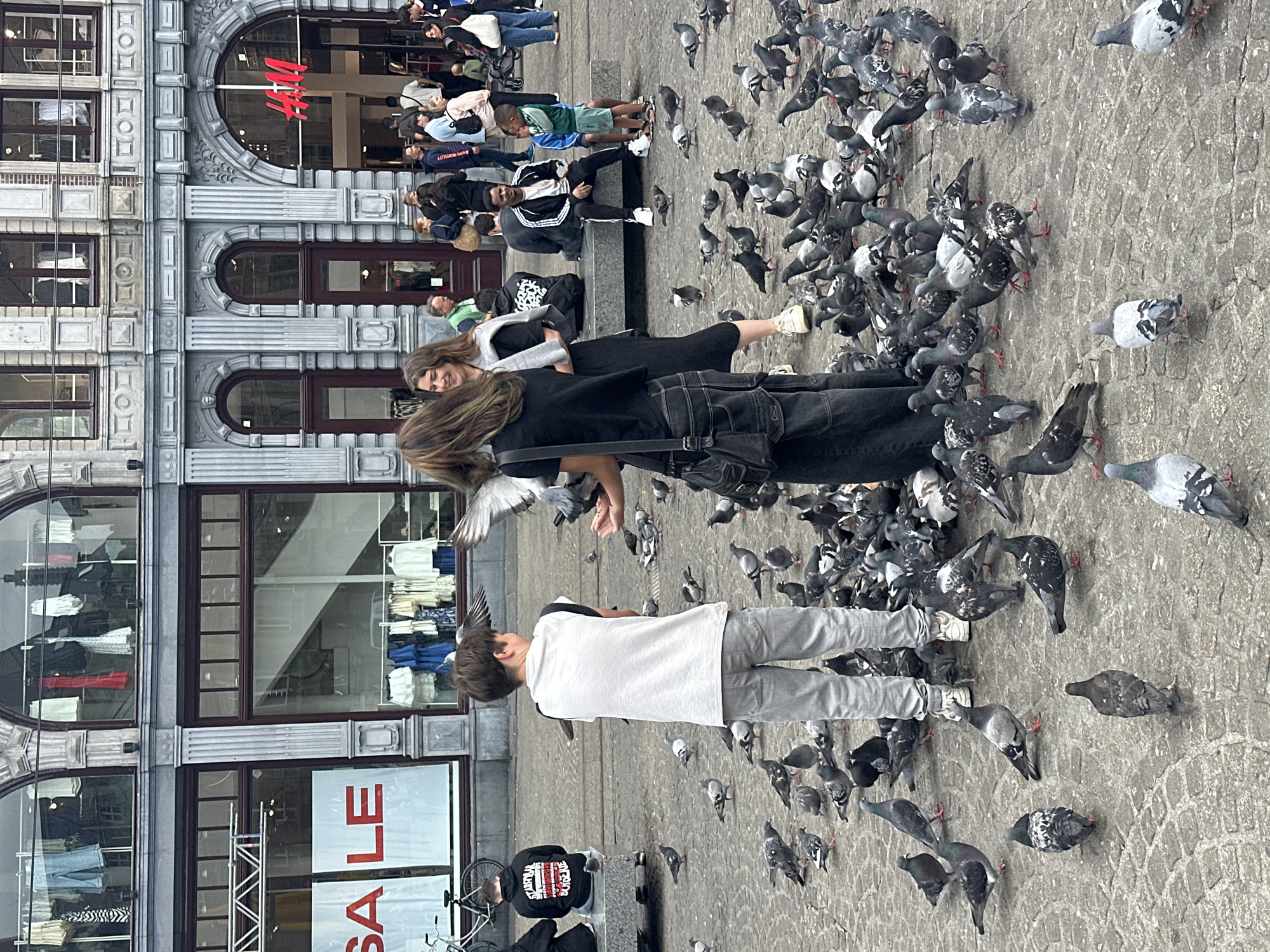 This image depicts a public square in Amsterdam, Netherlands, with the H&M store and another retail establishment visible in the background. In the foreground, a young boy in a white t-shirt and grey pants is surrounded by numerous pigeons. He appears to be interacting with the birds, possibly feeding them. To his right, a woman in a black outfit is also near the pigeons, with her hands held out. Several other people are scattered throughout the square, some observing the scene and others walking by. The ground is paved with cobblestones, typical of many European cities. The architecture in the background suggests a historical urban setting.