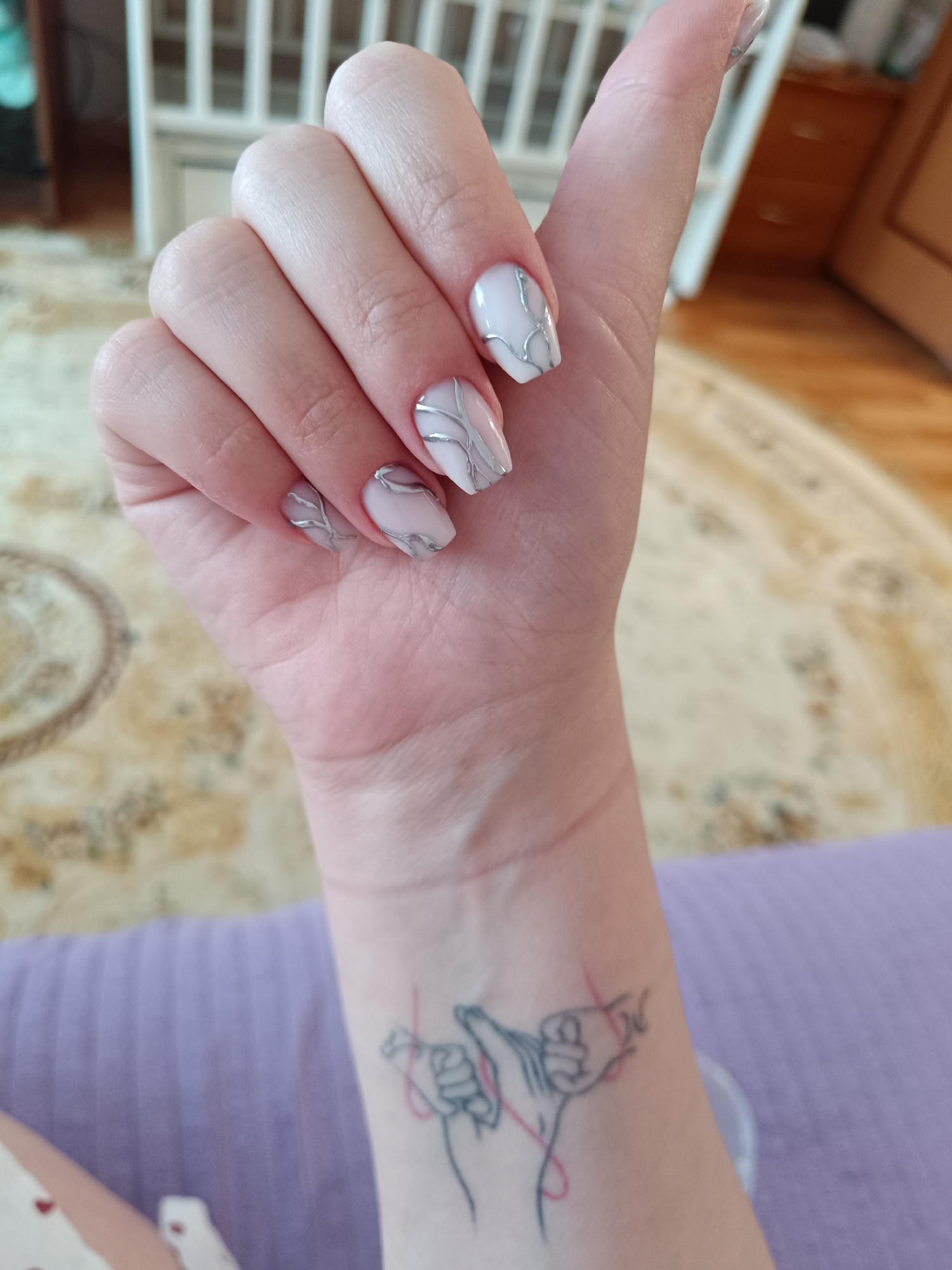 A close-up shows a fair-skinned human hand with manicured nails. The nail polish is light pink with a silver, vein-like design. On the wrist is a black line tattoo of two hands with interwoven red string. The background is blurred, but appears to include wooden furniture and a patterned carpet. There is no information to suggest the image location is Tobolsk, Russia.