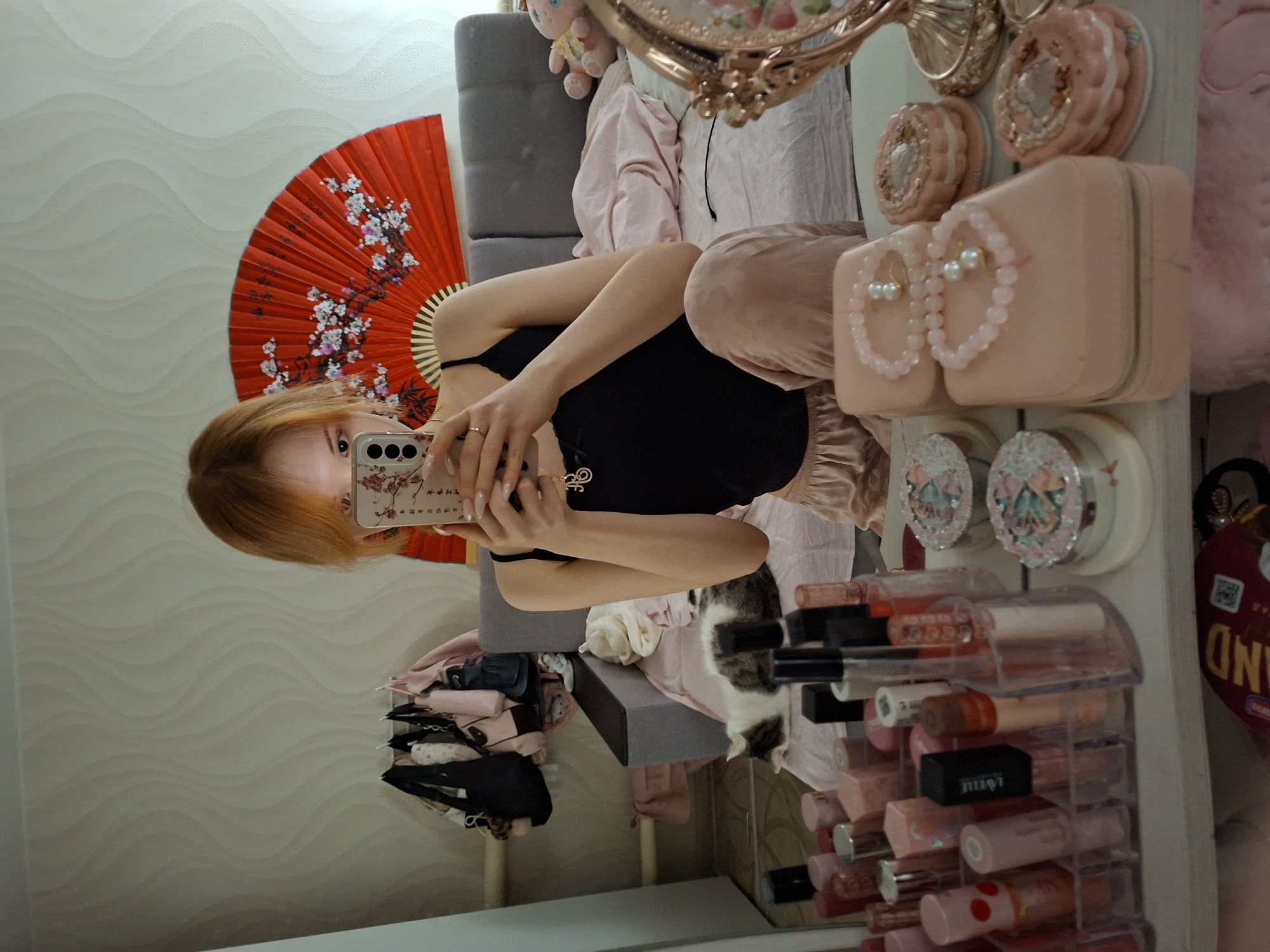 The image, correctly rotated, captures a person taking a mirror selfie in what appears to be a personal room, likely a bedroom or dressing area. The subject, visible from the chest up, has reddish-brown hair and is wearing a black top and light-colored (possibly pinkish) ruffled pants. They hold a smartphone with a white and pink floral-patterned case, which partially covers their face, revealing only their eyes and upper nose.
To the left, a vibrant red hand fan decorated with white and pink cherry blossom designs stands out against a light-colored wall with a subtle wavy pattern. Behind the person, part of a gray upholstered chair or sofa is visible.
The right side of the mirror reflection showcases a collection of decorative items and cosmetics. These include a vintage-style ornate gold mirror with pink accents, several pink, round, macaron-shaped containers, and rectangular pink cases. One of the pink cases is adorned with a pair of pearl-like pink beaded bracelets. A clear acrylic organizer is filled with various lip glosses and lipsticks. Further into the background, a small pink plush toy peeks out, and a tabby and white cat is seen resting on a pink blanket or clothing near the gray chair. Assorted bags, some pink and one black, along with folded garments, are also present.
Visible text includes delicate Chinese characters on the smartphone's floral case and the letters "OND" on a red heart-shaped item. The scene suggests a personalized, feminine, and well-accessorized space.