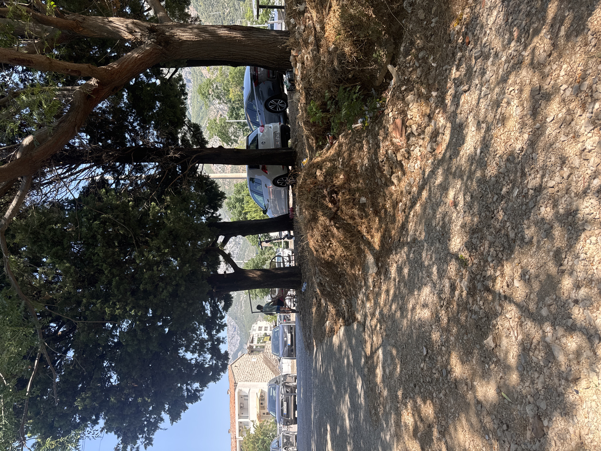 In Sutomore, Montenegro, a parking lot is bordered by trees and a hilly landscape. In the foreground, the ground is covered in gravel and dry vegetation. Several cars are parked in the lot, including a dark grey SUV with license plate "BD 2643 HD", a dark grey sedan with license plate "PG F481 EK", and a white Volkswagen Golf. Behind the white Golf, a dark grey sedan is parked. A person in a green shirt and blue shorts is walking near a metal structure, possibly a bench, with a scooter nearby. Bottles are scattered on the ground. In the background, buildings and mountains are visible under a clear blue sky.