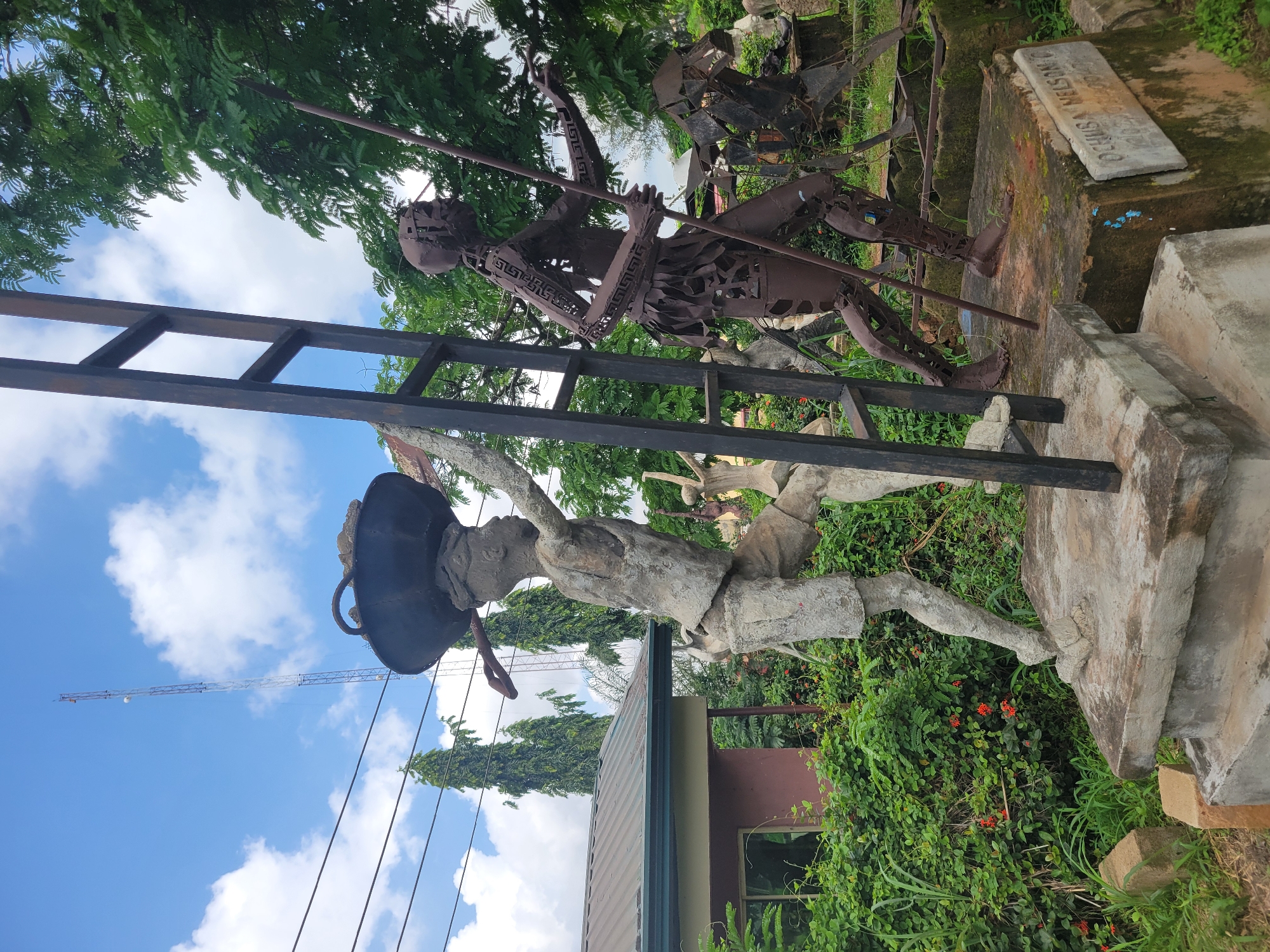 The media file depicts an outdoor scene in Egbagu, Nigeria, featuring several sculptures. In the foreground, a sculpture of a person made of concrete is climbing a dark metal ladder. This figure is wearing a large, dark hat that resembles a pot and is dressed in shorts and a shirt. Behind this figure, a sculpture of another person, made of dark, rusted metal with intricate cutouts, is also present. This metal figure is holding a long pole and appears to be reaching upwards, possibly mimicking a climbing or reaching action. The figures are positioned in an outdoor setting with lush green vegetation and trees surrounding them. The sky above is blue with scattered white clouds. In the background, a tall tower-like structure, possibly a communication tower or a crane, is visible against the sky. A stone plaque is mounted on a concrete base near the metal sculpture, with some visible text that appears to be "DENISA NELSON". The overall impression is that of an outdoor art installation in a garden or park-like environment. The lighting suggests it is daytime, and the weather appears clear.