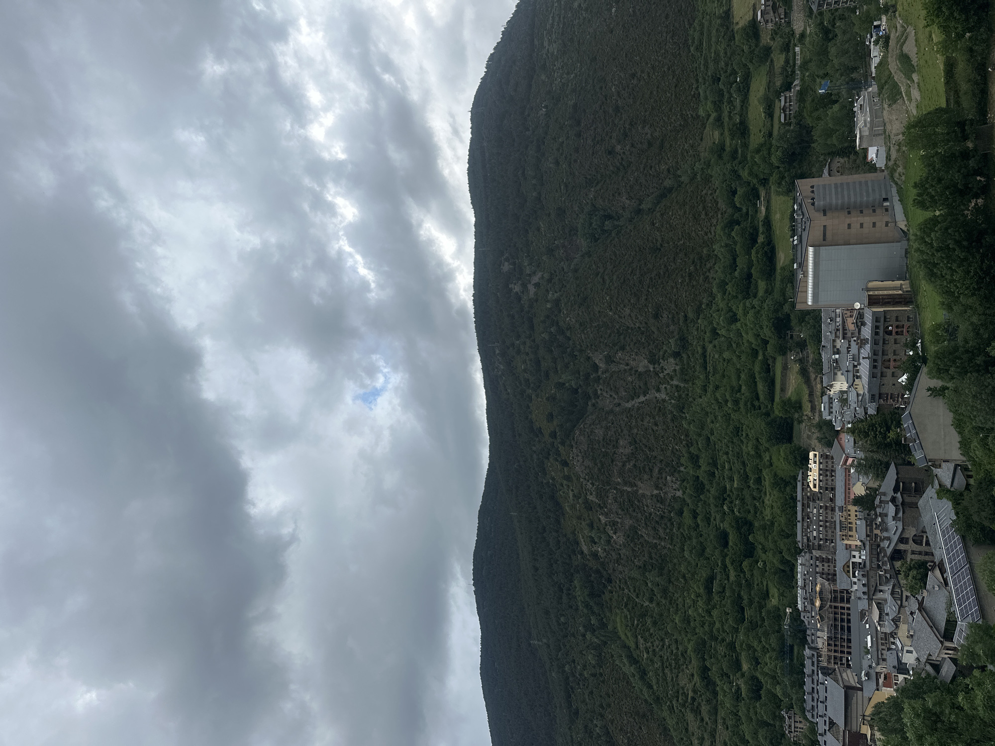 The image, when viewed in landscape orientation, captures a panoramic scene of Encamp, Andorra, nestled within a mountainous valley under an overcast sky. The upper left portion of the frame is dominated by a dense layer of grey, textured clouds, with a small patch of brighter blue sky visible, suggesting a break in the cloud cover.
A large, steep mountain, heavily forested with dark green trees, rises prominently on the right side, its rugged, tree-covered slope stretching from the valley floor upwards towards the cloud line. The mountain's peak is not fully visible, but its vast extent underlines the dramatic topography of the region.
At the bottom of the image, hugging the base of the mountain, lies a significant portion of the town. Numerous buildings of varying sizes and architectural styles are visible, characterized by predominantly grey and brown roofs, many of which are equipped with solar panels. The structures range from multi-story residential or commercial buildings to smaller dwellings, integrated closely with the natural landscape. Green foliage from trees and bushes is interspersed throughout the urban area and blends into the denser forest covering the mountain.
The time of day appears to be daytime, likely mid-morning or afternoon, with diffused light indicative of the cloudy conditions. There are no people visible, and no specific activities are taking place. No discernible text can be read on any of the buildings or signs. The scene conveys the characteristic integration of urban development within a striking natural mountain environment.