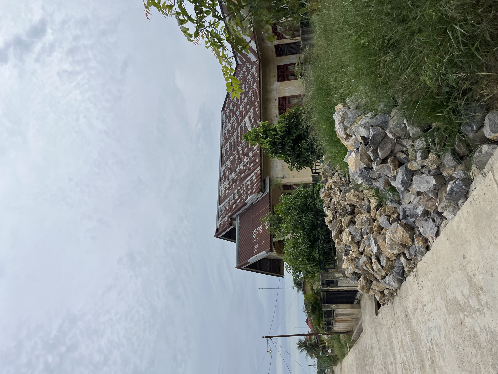This media file depicts an outdoor scene in Bundar, Indonesia, with a focus on a weathered building and its surroundings. The image is oriented vertically, showing a house with a rusty, corrugated metal roof and cream-colored walls. Several windows with red frames are visible on the side of the house. Lush green grass grows along the side of the building, and a pile of large, grey rocks is situated between the grass and a concrete path. Trees are also present, with some foliage hanging over the roof of the house. In the background, more buildings and palm trees are visible, suggesting a residential or village area. A power line with a single light fixture stretches across the frame. The sky is overcast with clouds, indicating potentially dull or rainy weather. The time of day appears to be daytime. No people are visible in the image, and no text is present. The overall impression is one of a somewhat neglected but functional living space in a tropical or subtropical environment.
