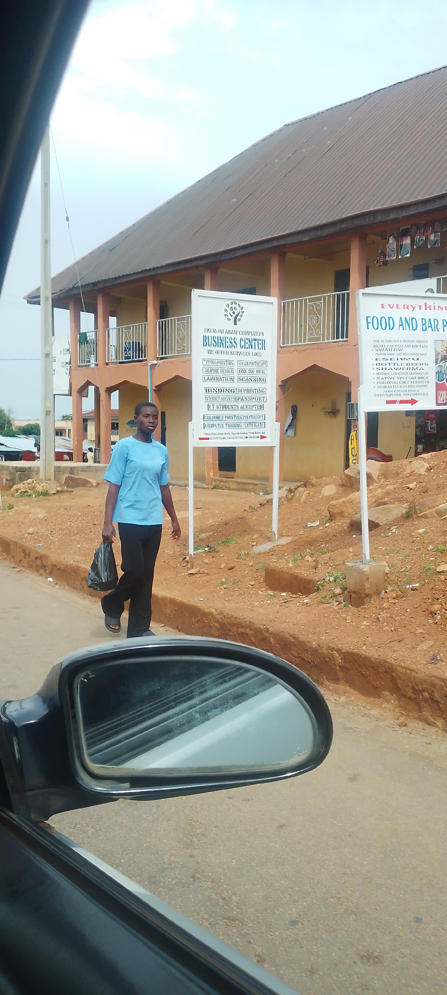 A young woman in a blue t-shirt and black pants walks on the side of a dusty road in Jos, Nigeria. She carries a black plastic bag in her left hand. She is walking past two white signs, one advertising "EKEMINI ABASI COMPUTERS BUSINESS CENTER" with services like typing, printing, and graphic design, and another, partially visible, that appears to be a food and bar advertisement. Behind the woman is a light-colored building with an orange trim and a corrugated iron roof. The area appears to be a commercial or residential district with some debris and vegetation on the roadside. A car's side mirror is visible in the foreground, reflecting the road and the sky.
