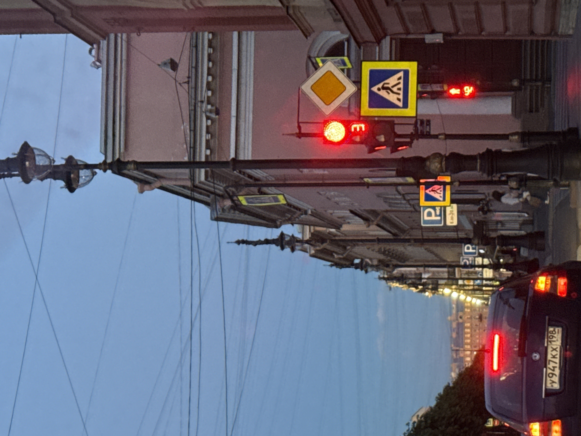 The image shows a street scene in Saint Petersburg, Russia, during twilight or early evening. A dark-colored car is in the foreground, facing away from the camera, with its brake lights illuminated. The car has a license plate reading "y947KX 198". A traffic light is visible, showing a red signal. Above the red signal, a digital display shows the number "3". A pedestrian crossing sign is mounted on a pole. In the background, there are ornate street lamps and buildings with classical architecture, some of which are lit. The sky is a deep blue, indicating the transition from day to night. The street is lined with trees on the left side. Several road signs are visible, including parking signs and a pedestrian crossing sign with a yellow square background and a blue pedestrian symbol. Another traffic light further down the street shows a red pedestrian signal with the number "42" displayed. The overall impression is a typical urban street at dusk, with elements of historic architecture characteristic of Saint Petersburg.