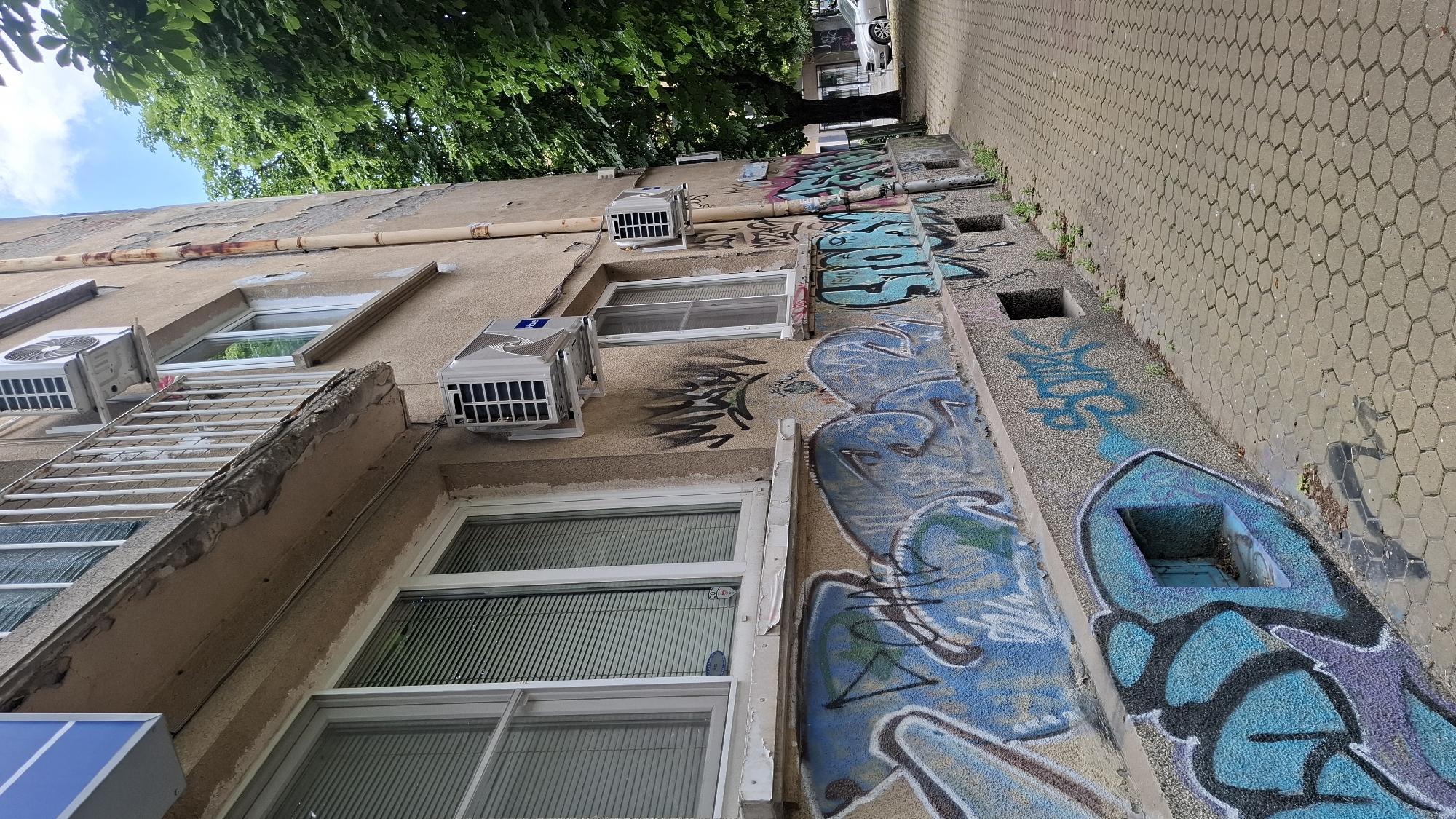The image shows a narrow sidewalk in Sofia, Bulgaria, running alongside a dilapidated building. The building's facade is covered in graffiti, with vibrant blue and black spray paint dominating the lower sections. Several air conditioning units are mounted on the exterior walls, and the balcony above the windows shows signs of wear and tear. The sidewalk is paved with hexagonal gray tiles, and a large green tree with dense foliage overhangs the scene from the right. In the distance, a silver car is parked on the street, and the sky above is partly cloudy. There are no people visible in the frame, and the overall impression is one of urban decay and neglect.