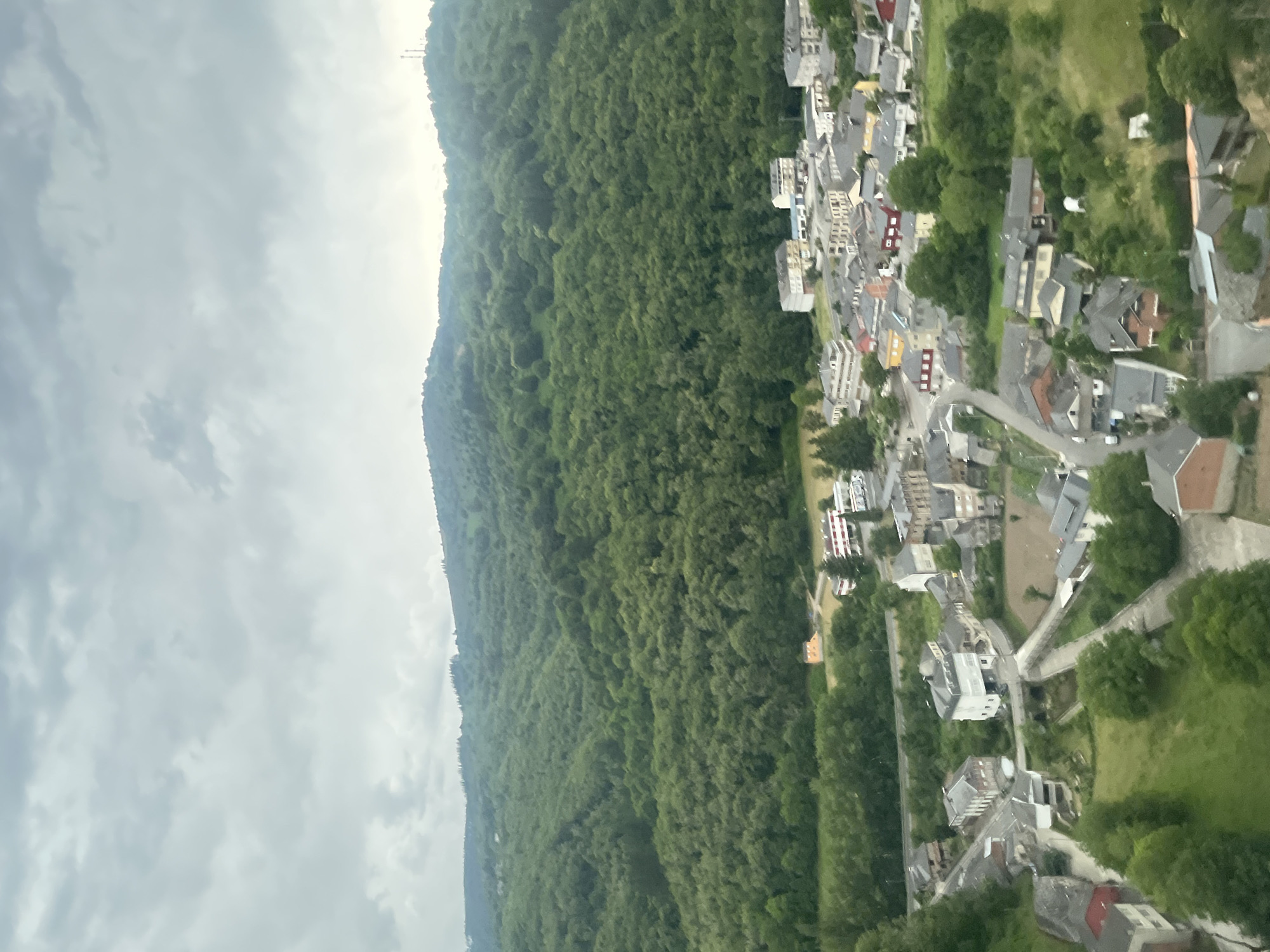 This is an aerial view of the town of As Nogais, Spain, nestled amidst a lush, green, hilly landscape under a cloudy sky. The town is a collection of buildings with red, gray, and white roofs, tightly packed together along winding roads. In the foreground, several houses with red roofs are visible. Further into the town, larger buildings, some with multiple stories, are clustered together. The town is surrounded by dense, vibrant green forests that extend up the rolling hills, with the trees appearing full and verdant. The sky is overcast with a mix of light and dark gray clouds. The overall scene depicts a rural town integrated into its natural environment.