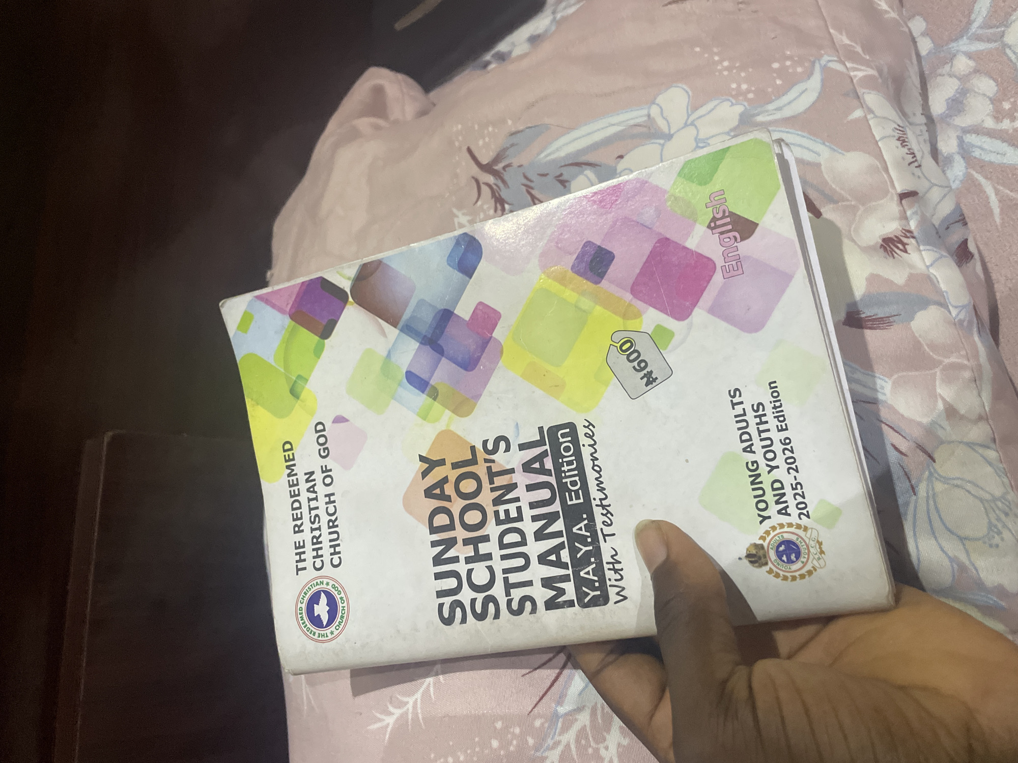 In Jalingo, Nigeria, a person's hand holds a "Sunday School Student's Manual" titled "Y.A.Y.A. Edition With Testimonies." The cover features a colorful abstract pattern of squares in shades of pink, purple, yellow, and green. At the top, the logo of "The Redeemed Christian Church of God" is visible, with the words "The Redeemed Christian Church of God" printed next to it. A price tag indicating "N600" is present on the lower right side of the cover. Below the title, another emblem is seen with "Young Adults and Youths 2025-2026 Edition" printed around it. The manual is resting on a pink surface, possibly a bed, with a floral pattern. The background is dark and out of focus, suggesting an indoor setting.