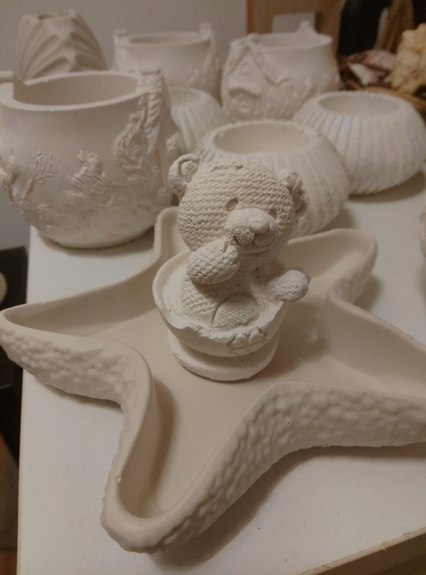 The image displays a collection of unpainted ceramic crafts, predominantly in the shape of pots and decorative items, set against a neutral, light-colored surface. In the foreground, a white ceramic starfish-shaped dish holds a small, textured teddy bear figurine sitting inside a round base. The teddy bear has detailed paws and a textured body, with its arms raised and holding a small object. Behind the starfish dish, several other ceramic pieces are visible, including a large, ornate pot with raised detailing resembling sea life and foliage, and a round pot with a ribbed texture. Further back, more ceramic items are present, some appearing to be small houses or planters with embossed patterns. The overall scene suggests an art studio or craft workshop environment. The image does not provide any specific information about the location in the city of Wedel, Germany.