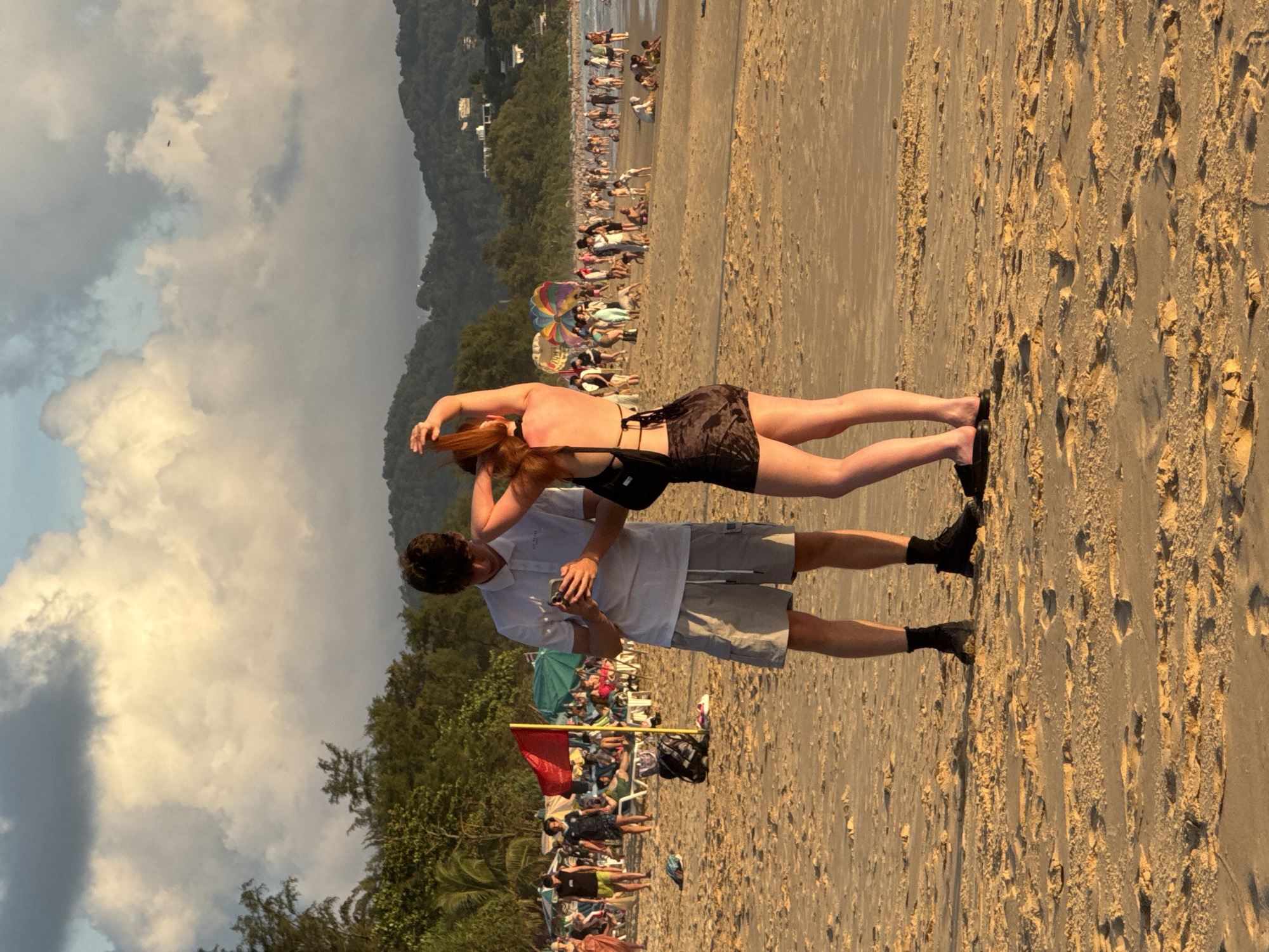 This image depicts a beach scene in Karon, Thailand, during what appears to be late afternoon or early evening. In the foreground, a young man wearing a white polo shirt and gray shorts is looking at his phone. Beside him, a young woman with red hair, wearing a black dress, is adjusting her hair. Both are standing on the sandy beach, with footprints visible in the sand.
In the midground, the beach is populated with many people. Umbrellas and lounge chairs are set up, and individuals are seen walking, sitting, and interacting. A red flag is planted in the sand near a cluster of umbrellas. Further in the background, the coastline is lined with lush green hills dotted with buildings. The sky above is filled with dramatic clouds, illuminated by the setting sun, casting a warm glow over the scene. The overall atmosphere is that of a busy tourist beach.