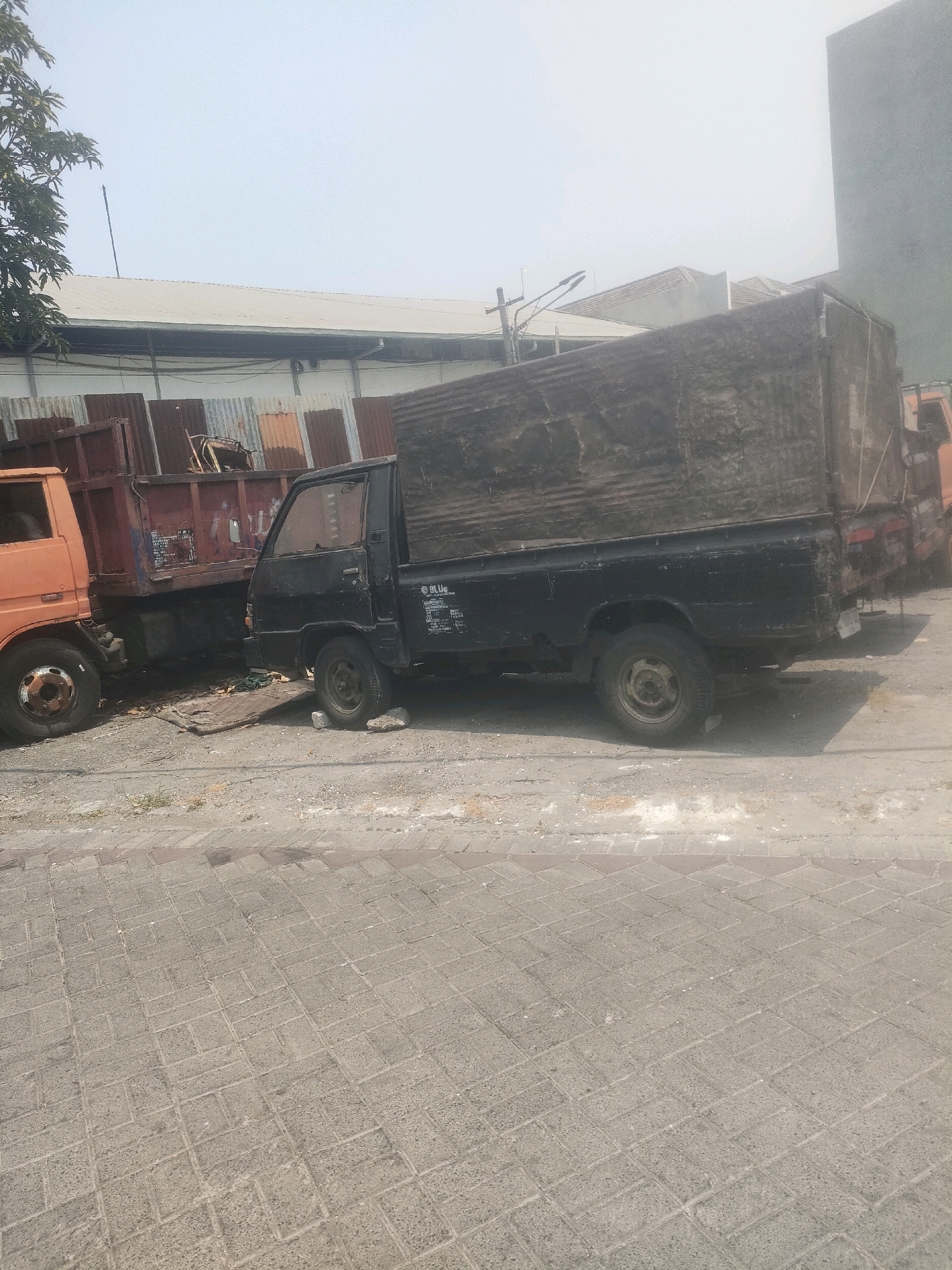 The image shows an outdoor scene in Surabaya, Indonesia. In the foreground, there is a paved area with interlocking brick-like stones. Behind this, a dirt and gravel area separates the pavement from two trucks. The truck on the left is an orange dump truck. To its right, a black pickup truck with a canvas-covered bed is parked. The background features a corrugated metal fence and a building with a light-colored roof. The sky is overcast, and there is some foliage visible to the far left. The scene appears to be in an industrial or commercial area, possibly a parking lot or storage yard. No people are visible in the image.