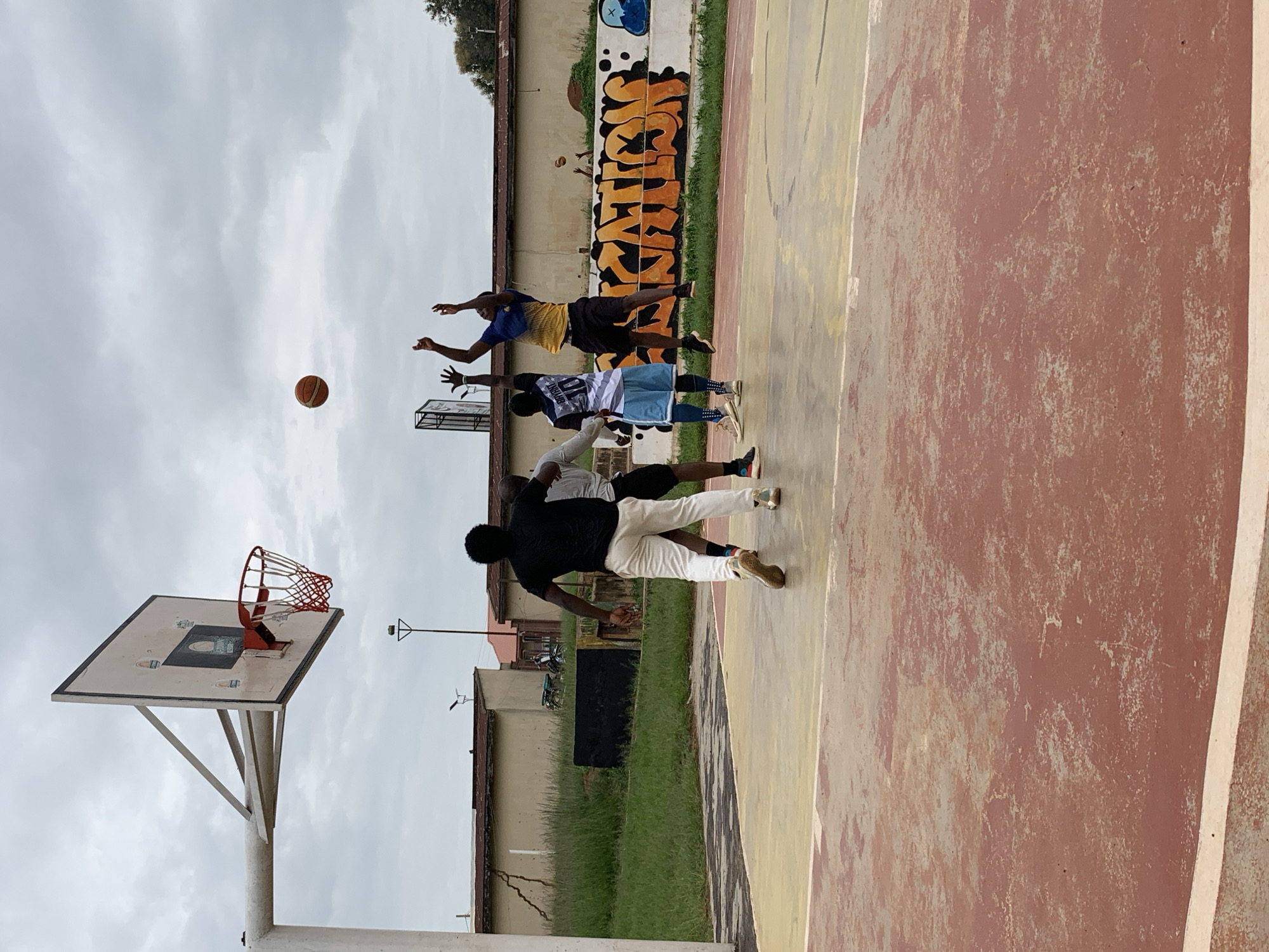 The image depicts a basketball game on an outdoor court located in Jos, Nigeria. Four young men are actively engaged in the game. One player in a blue and yellow jersey is jumping and reaching for the ball, while another player in a black t-shirt and white pants is positioned nearby, observing the play. Two other players, one in a white and blue jersey with the number "10" and the other in a grey t-shirt, are also on the court. The basketball is airborne in the center of the frame. The court itself is a faded red and yellow surface. In the background, there are buildings with light-colored walls, a patch of green grass, and a wall adorned with graffiti featuring large orange lettering. The sky is overcast with grey clouds.
