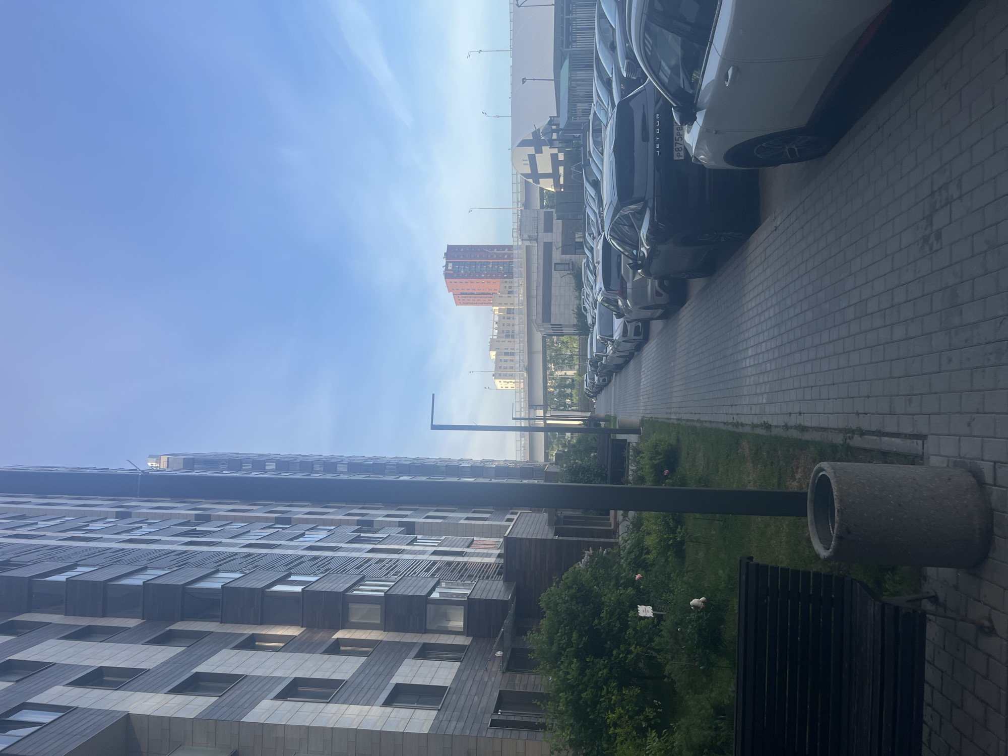 This image captures a modern urban landscape, likely a residential area in Moscow, Russia. The scene is oriented vertically.
On the left side, a tall, contemporary apartment building dominates the foreground, featuring a patterned facade combining dark grey and lighter panels with numerous windows. Green bushes and grass line the base of the building, separating it from a paved walkway.
To the right, a row of parked cars, predominantly dark-colored SUVs or crossovers, extends into the distance along a paved surface made of light-colored bricks or pavers. One of the dark SUVs prominently displays the brand name "JETOUR" on its rear and a Russian license plate with the characters "P875PA". A tall, dark lamppost stands near the center of the frame, extending above the foreground vegetation. In the lower right, a large concrete pipe or planter is visible next to a dark, slatted bench.
In the mid-ground and background, several other buildings can be seen under a bright, hazy blue sky. A distinctive reddish-orange high-rise building stands out against the horizon, along with what appears to be an elevated road or bridge structure.
The clear, bright sky suggests it is daytime, possibly morning or afternoon. The scene is static, with no specific activity or event taking place, depicting an everyday view of a modern city street.