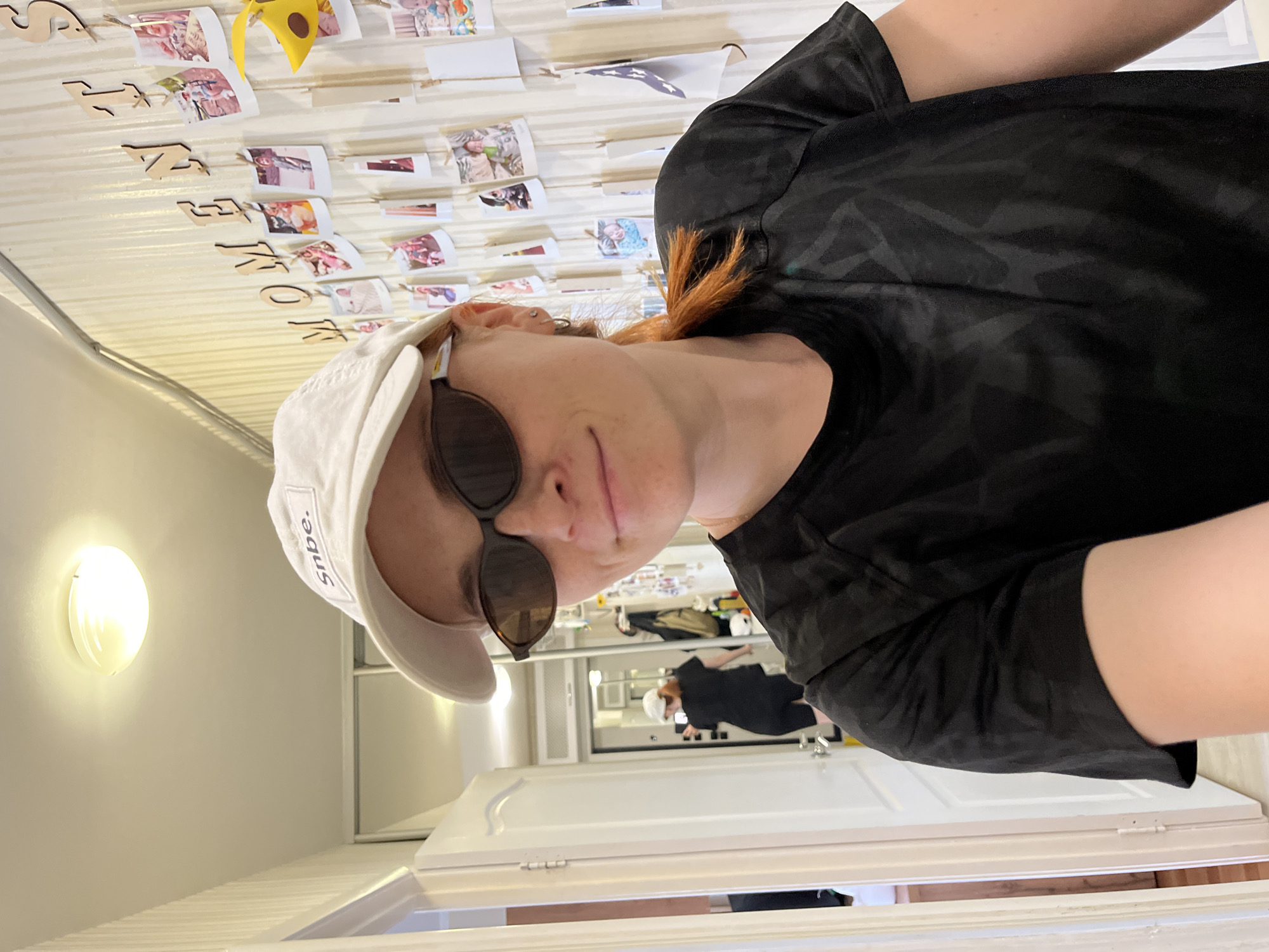 A person with reddish-brown hair, wearing a black t-shirt, sunglasses, and a white baseball cap, looks towards the camera. The cap has the word "snbe." visible on it. The person is in an indoor setting, possibly a room or hallway, with white walls and trim. In the background, there are several white doors. The person appears to be looking slightly to the side with a neutral expression. The lighting suggests it could be daytime. There is a string of wooden letters and several small printed photos hanging on a wall in the background. A yellow object, possibly a decoration, is also visible among the photos. The image is taken from a slightly low angle, looking up at the person. There are no clear indicators of the weather or specific time of day beyond the interior lighting.