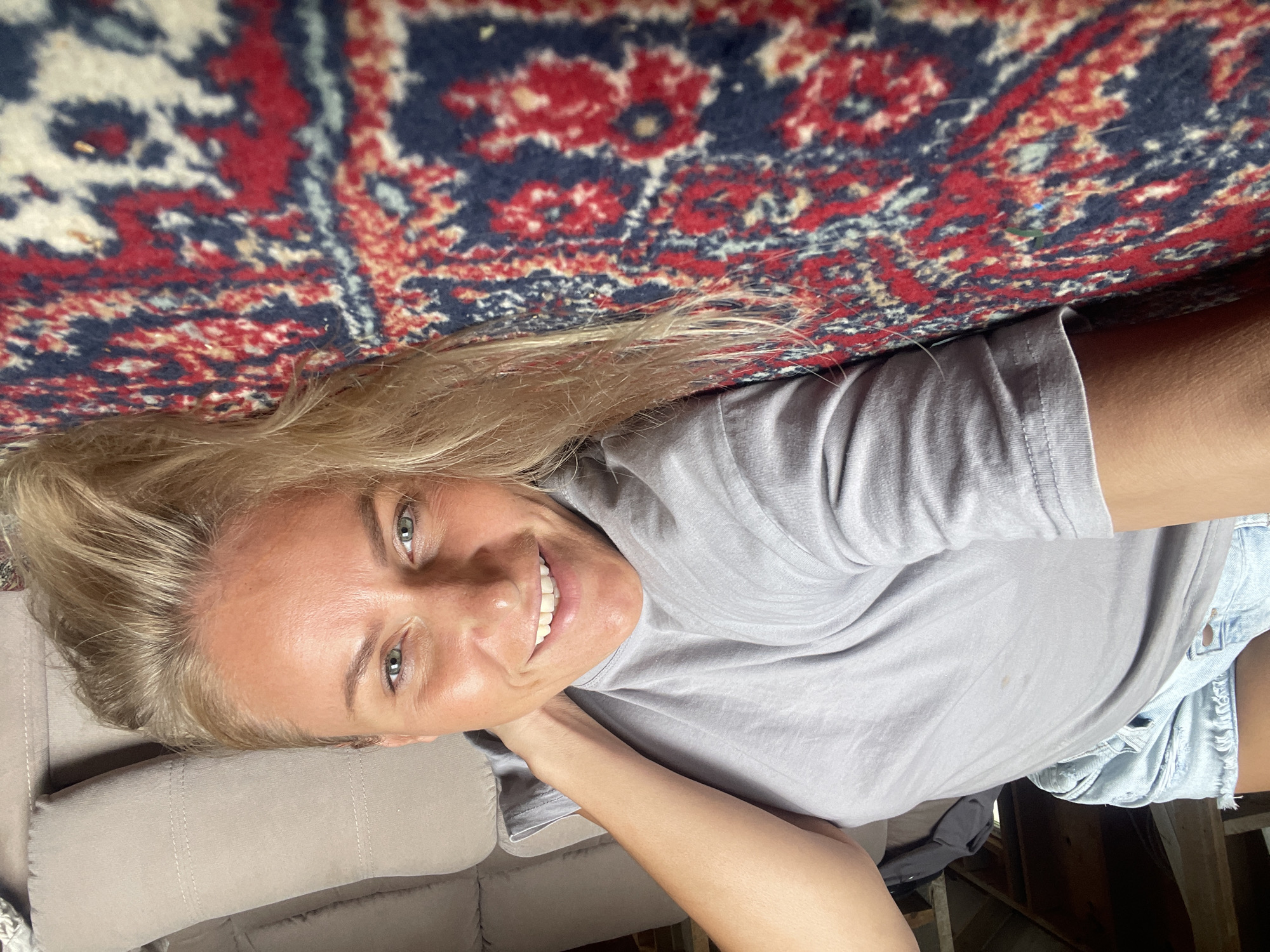 A woman with blonde hair is lying down, smiling at the camera. She is wearing a grey t-shirt and denim shorts. Her head rests on a beige-colored sofa. A patterned rug with red, navy, and cream colors is above her. Parts of a wooden structure, possibly a ceiling or floor, are visible in the lower right corner. The location is unidentifiable as Baranovka, Russia.