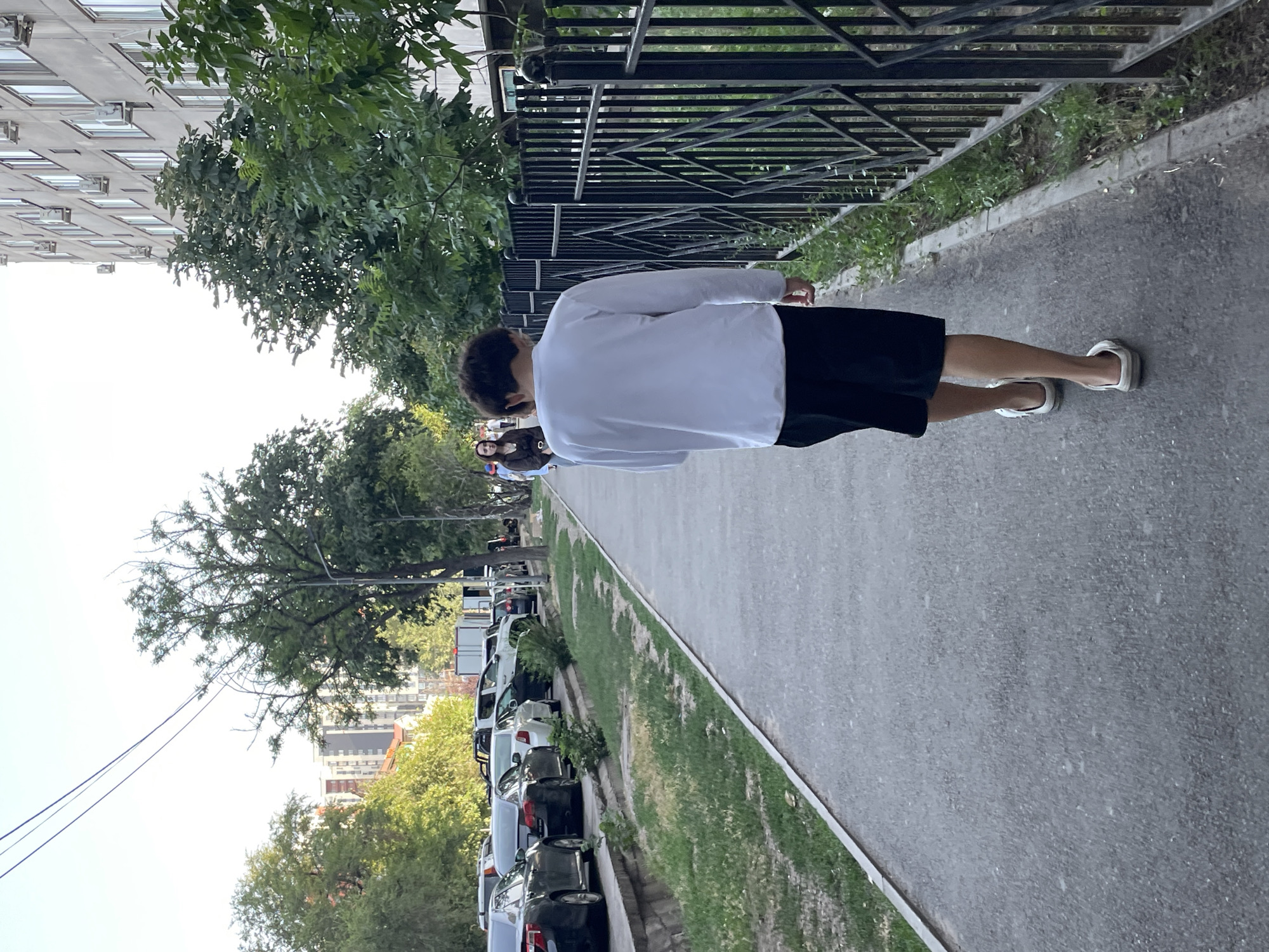 A young man walks away from the camera on a paved sidewalk in Almaty, Kazakhstan. He is wearing a white long-sleeved shirt and black shorts. He is also wearing white sandals. The sidewalk is lined with a black metal fence on the right and a grassy area with parked cars on the left. In the background, there are trees and apartment buildings. The sky is clear and bright.
