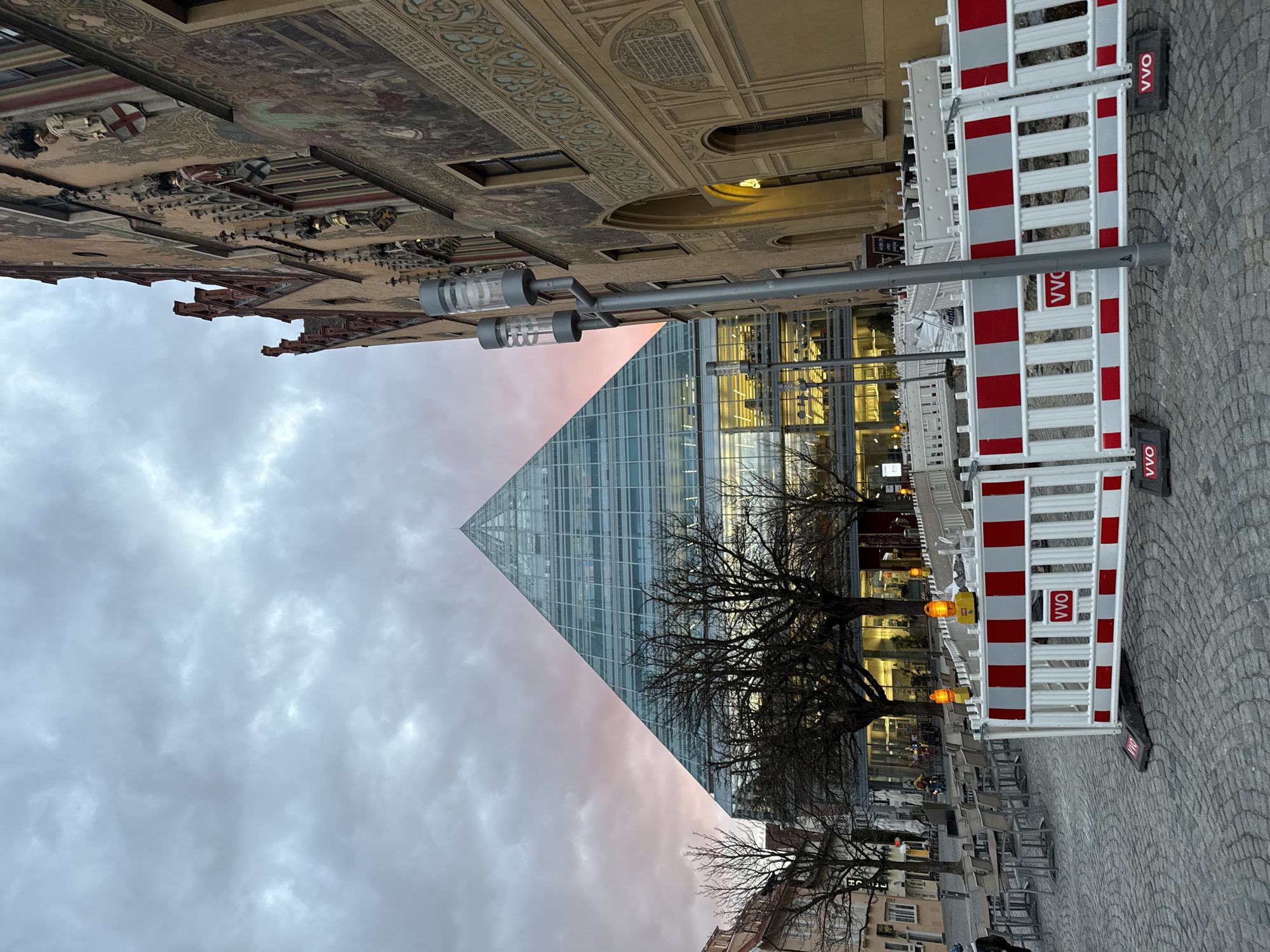 This image captures an urban scene at dusk in Ulm, Germany, specifically featuring the Ulm Public Library (Stadtbibliothek Ulm) and the Ulm City Hall (Rathaus).
The foreground is a light grey cobblestone plaza. A series of white and red striped plastic construction barriers, marked with "VVO" in red text and equipped with yellow flashing warning lights, delineate an area on the right. To the left of these barriers, numerous empty outdoor cafe tables and chairs, made of grey metal, are arranged on the cobblestones. A prominent grey, twin-fixture streetlight stands on the right side of the frame. A lone individual, dressed in dark attire, is partially visible on the far left edge, near the cafe tables.
In the midground, several bare deciduous trees are silhouetted against the backdrop of the Ulm Public Library. This modern building features a distinctive pyramid-shaped glass and steel construction, with its interior floors brightly illuminated, revealing an open-plan layout. Indistinct figures are visible through the library's glass facade, and a person on a bicycle or scooter is visible in the distance near the cafe area.
The right side of the background is dominated by the historic Ulm City Hall. This ornate building displays a light brown facade adorned with intricate Gothic architectural details, including gables, sculpted figures with heraldic shields, and extensive murals or frescoes depicting historical scenes and decorative patterns. Arched windows and doorways are visible along its lower levels. On the far left background, several multi-story traditional European buildings with typical window patterns are present.
The sky is heavily overcast with dark grey clouds, exhibiting a distinct pinkish-red hue along the horizon, consistent with either sunrise or sunset.