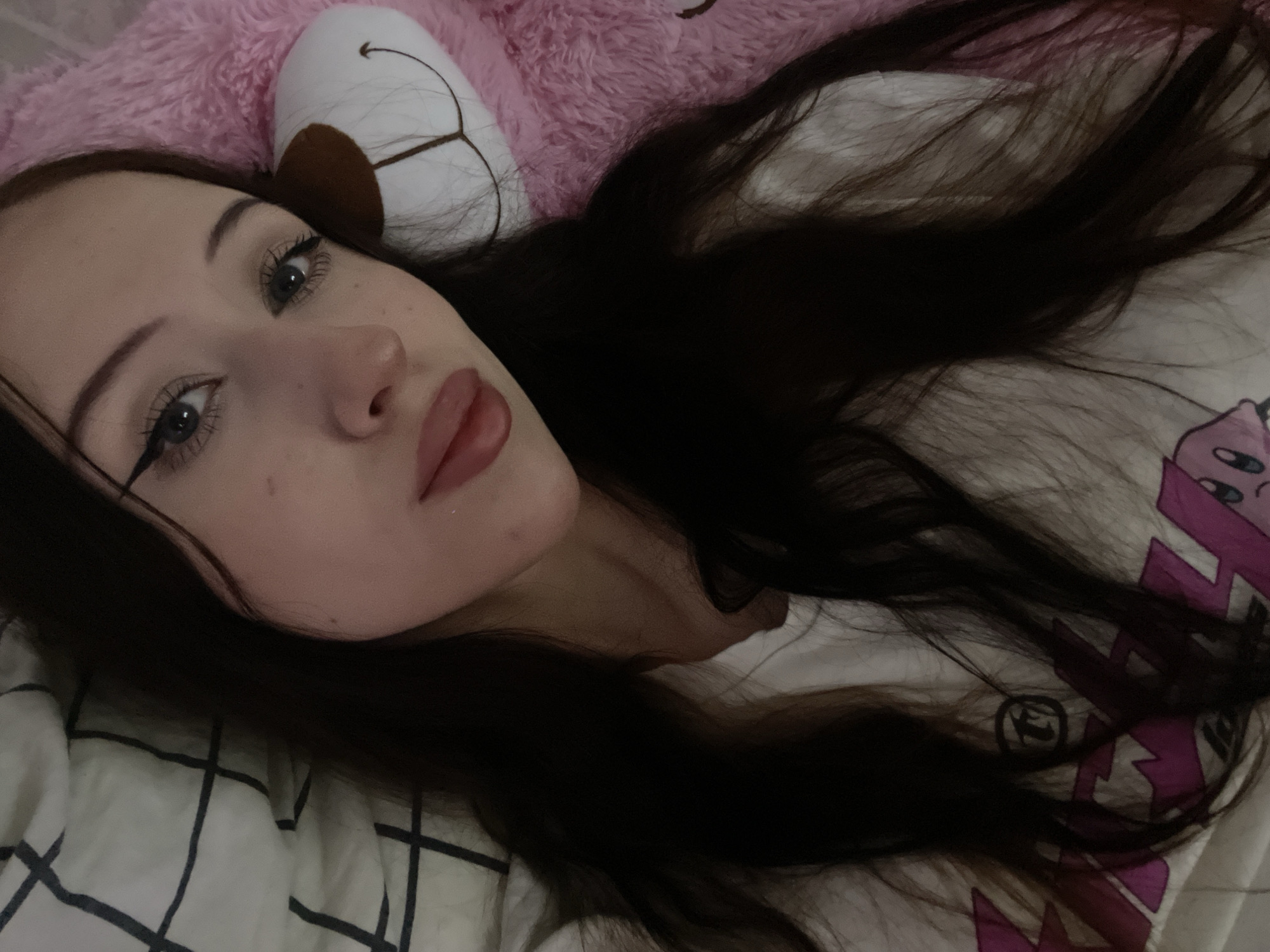 This is a close-up, high-angle, medium-shot media file of a young woman lying down in bed, looking at the camera. She has long, dark brown hair that is spread out around her. She is wearing makeup, including eyeliner and lip gloss. She has light skin and is lying on a white and black checkered sheet. Behind her head is a pink, fuzzy object that appears to be a stuffed animal or pillow. A portion of a white t-shirt with a purple graphic design is visible on her right side. The lighting suggests it is either nighttime or the room is dimly lit. Her expression is neutral or slightly curious. There is no discernible weather or time of day information beyond the indoor setting. No text is visible in the image. The location context provided is Votkinsk, Russia.