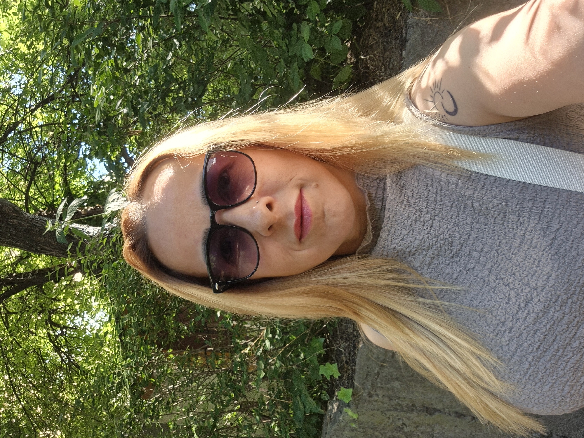 A blonde woman with sunglasses and a grey top stands in front of lush green foliage and a stone wall. She has a tattoo on her right arm. The scene is outdoors, suggesting a park or natural area within a city.