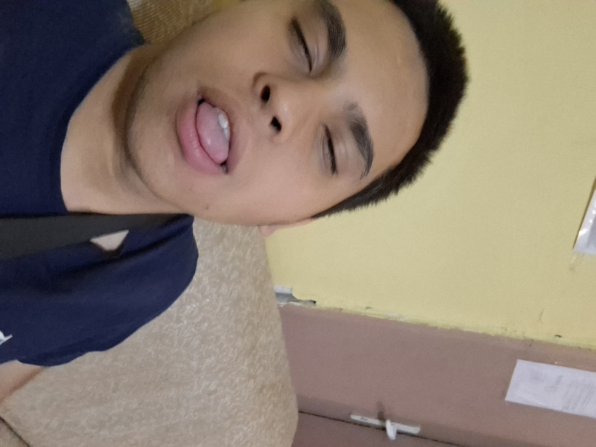 A young man is shown lying down, appearing to be asleep or unconscious, with his eyes closed and mouth open, his tongue partially visible. He has short, dark hair and is wearing a dark blue t-shirt. The image is significantly tilted, with the man's head positioned towards the lower right. He rests on a light brown, textured surface, possibly upholstery from a sofa or bed. The background features a wall painted in two sections: the upper portion is light yellow, and the lower part is a muted pinkish-brown, separated by a horizontal line. Scattered in the background are a few small, white, indistinct objects, possibly paper or tape affixed to the wall. The scene suggests an indoor environment, likely a room. There is no legible text visible. The time of day and specific location cues are not discernible from the image.