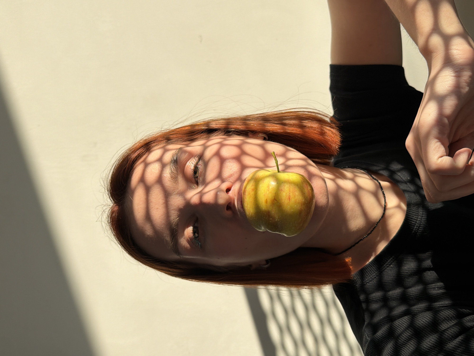 An indoor, close-up portrait depicts a female individual in the city of Chaikovsky, Russia. She has reddish-brown, shoulder-length hair and is wearing a black ribbed short-sleeved top and a thin black beaded necklace. A yellow and red apple is positioned horizontally in her mouth, covering her lips. Strong, directional sunlight casts prominent, repeated hexagonal grid-like shadows across her face, neck, upper torso, and the plain, light-colored wall behind her. Her eyes are open, looking directly forward, with some shadow obscuring their upper portions. Her left hand is visible in the lower right quadrant of the image, resting on an unidentifiable surface, also marked by the grid shadow pattern.