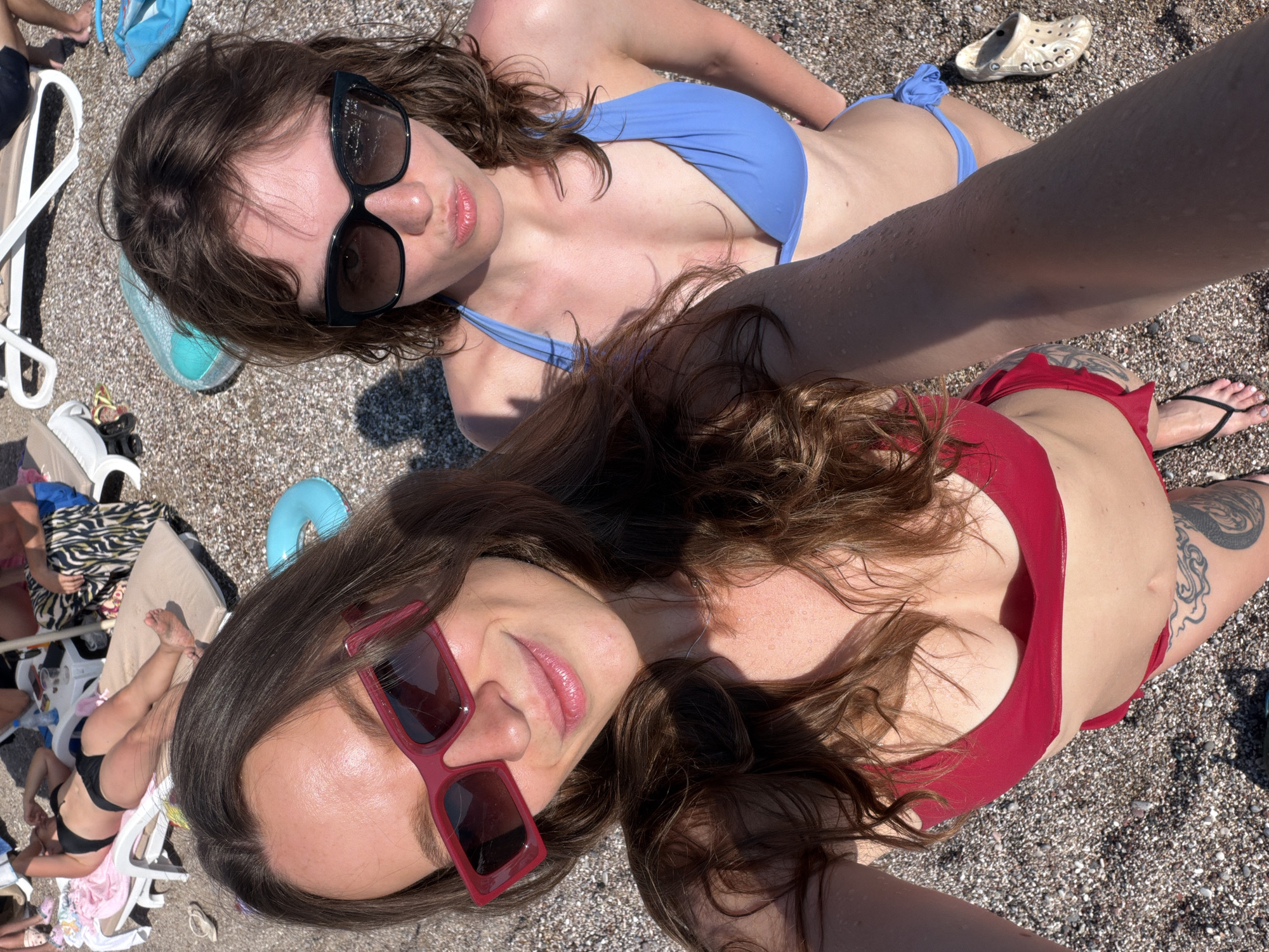 Two young women are posing for a selfie on a pebble beach in Sutomore, Montenegro. The woman in the foreground, on the left, is wearing a red bikini and red-framed sunglasses. She has long, brown, wavy hair and is smiling. The woman on the right is wearing a blue bikini and large, dark sunglasses. She has shorter, brown, wavy hair and is making a duck face. In the background, other beachgoers are visible lounging on chairs and walking. Beach accessories such as towels, a lifebuoy, and a pair of Crocs are scattered around. The scene appears to be a sunny day at the beach.
