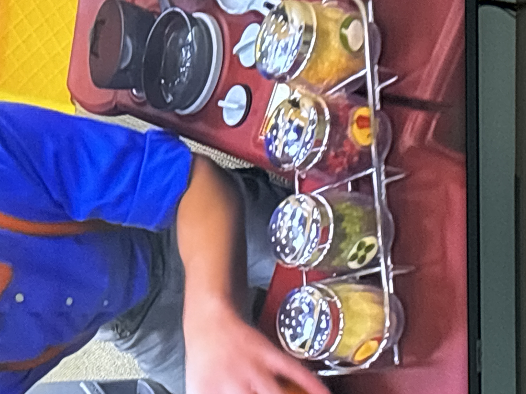 The image shows a close-up of a person's arm and torso wearing a blue shirt with orange trim, suggesting a uniform or costume. In the foreground, there is a rack holding four clear spice jars filled with colorful contents: yellow, green, red, and yellow again. The spice jars have silver lids with holes. Behind the spice jars is a red toy stove with black pots. The background is blurred, with hints of yellow and grey. The scene appears to be indoors, possibly a playroom or a set. There is no indication of the city of Dubai or the United Arab Emirates in this image.