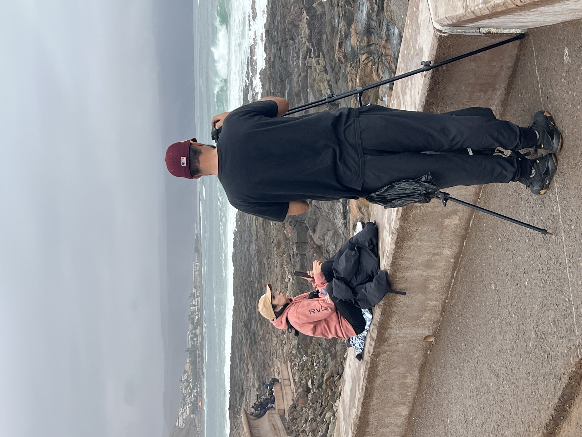 A man in a maroon baseball cap and black t-shirt is standing with his back to the camera, operating a video camera mounted on a tripod. He is wearing black athletic pants and black sneakers. In the foreground, a woman in a pink hoodie and tan baseball cap is sitting on a concrete ledge, holding a phone in her hand. A black jacket and a blue and white patterned bag are on the ledge next to her. The background shows a rocky coastline with waves crashing on the shore. In the distance, a town with white buildings is visible along a hillside. The sky is overcast and gray. This scene is consistent with the coastal areas of Arica, Chile, known for its rugged beaches and ocean views.