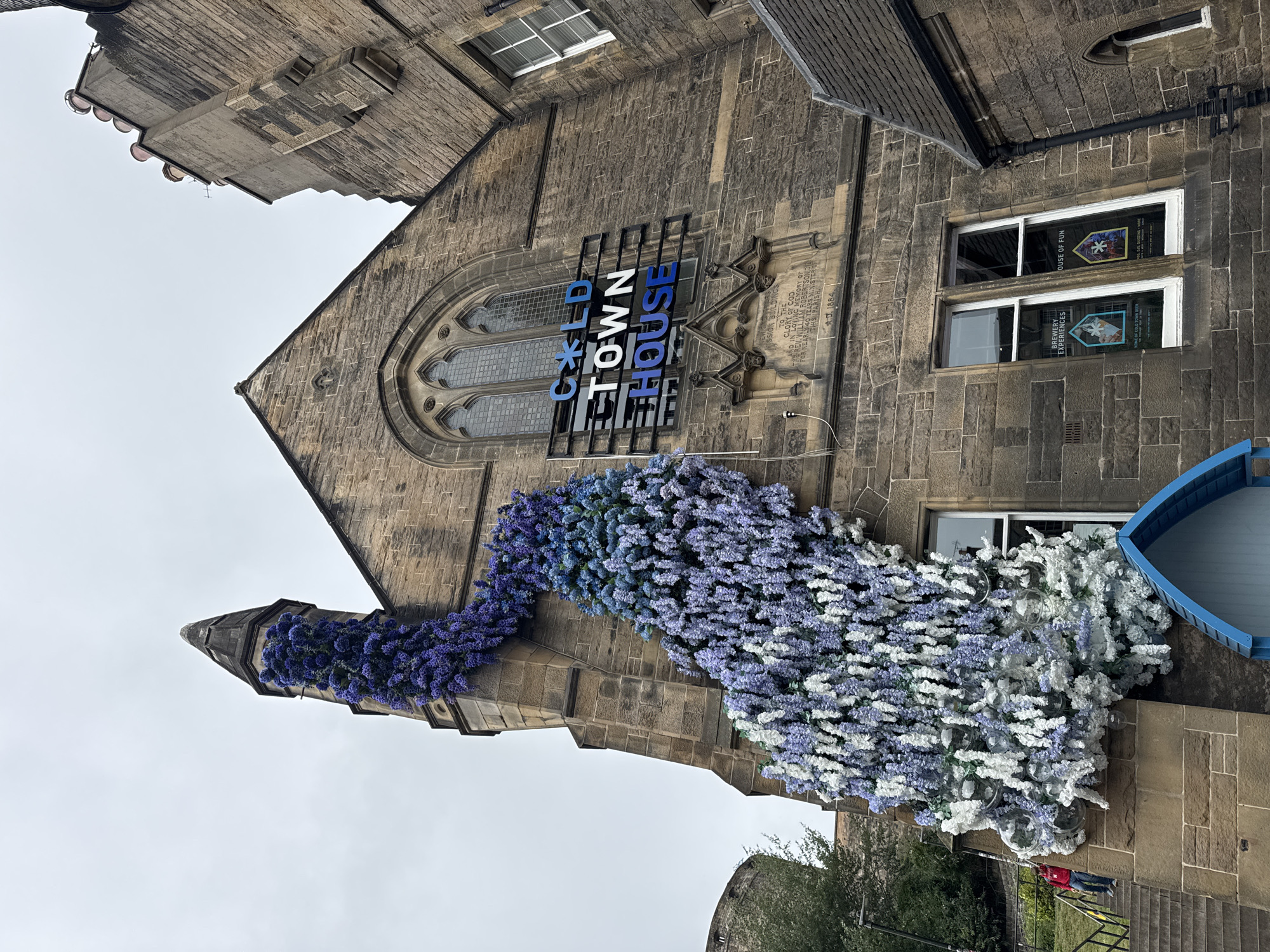 A stone building, possibly a church or historic structure, is adorned with a large, cascading floral installation. The installation features shades of purple, blue, and white flowers, artfully arranged to appear as if they are growing down the side of the building and over a prominent spire. The building itself has a gothic architectural style, with arched windows and stone detailing. A sign that reads "COLD TOWN HOUSE" is mounted on the facade. In the lower left of the image, a person in a red jacket is walking up a set of stone stairs. The sky is overcast. The building is situated in Edinburgh, Scotland, as indicated by the "COLD TOWN HOUSE" sign, which is a well-known establishment in the city.